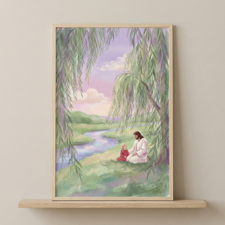 A soft-toned watercolor illustration depicts Jesus in white robes sitting on a grassy bank beside a young girl in a red dress. A winding blue river flows through rolling green hills under a pastel sunset sky of lavender and gold. Long, delicate branches of a weeping willow tree frame the peaceful scene from above. The artwork is presented in a light wood frame resting on a matching wooden shelf against a neutral-colored wall.
