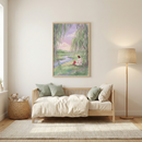 A minimalist child's room features a framed illustration on a white wall above a light wood daybed. The artwork depicts Jesus sitting on a grassy riverbank under a weeping willow, talking with a small child in a red dress during a pastel sunset. The daybed is dressed with neutral bedding and soft green pillows. To the left sits a woven basket filled with plush toys, while a simple floor lamp stands to the right. The room has light wood floors, a textured rug, and soft natural light.