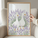 A vertical watercolor-style illustration features two white ducks with orange beaks and feet, standing in a field of tall purple lavender flowers and green leaves. The duck on the left is positioned slightly in front of the other. The artwork is housed in a light-colored wood frame and is being held by a hand on the right side, with the frame resting against a plush, textured beige chair. The overall aesthetic is soft, whimsical, and gentle.