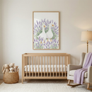 A bright and airy nursery features light-colored walls and a soft, textured white rug. In the center stands a light-toned wooden crib with a simple slatted design and a white mattress. Above the crib, a large vertical artwork is displayed in a matching wooden frame, depicting two white waterfowl standing amidst a dense field of tall, purple flowering herbs. 
