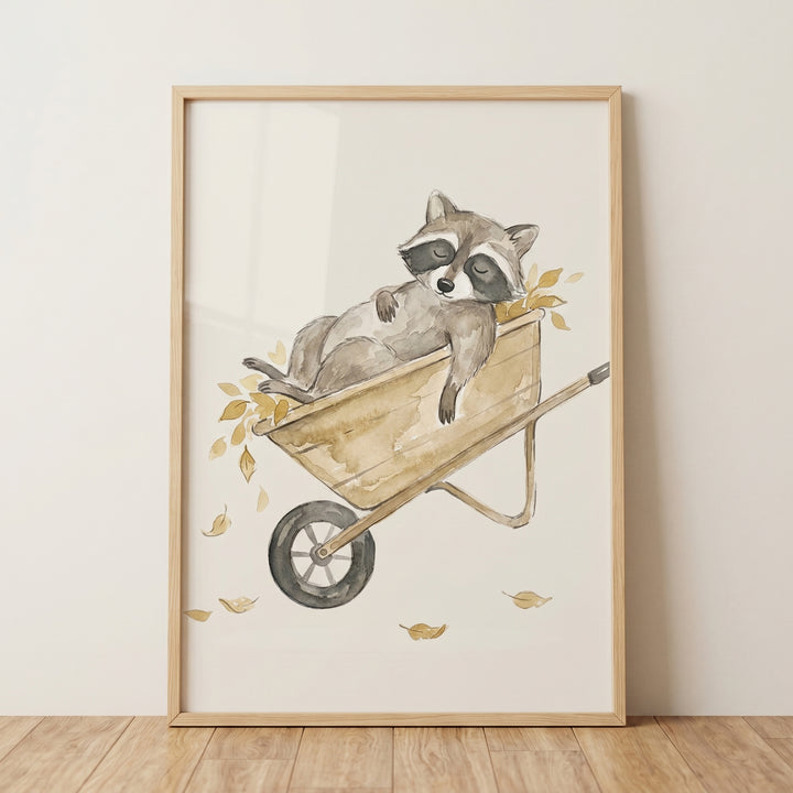 A sleeping raccoon rests in a cart full of gold leaves in this watercolor leaf wheelbarrow raccoon wall art.
