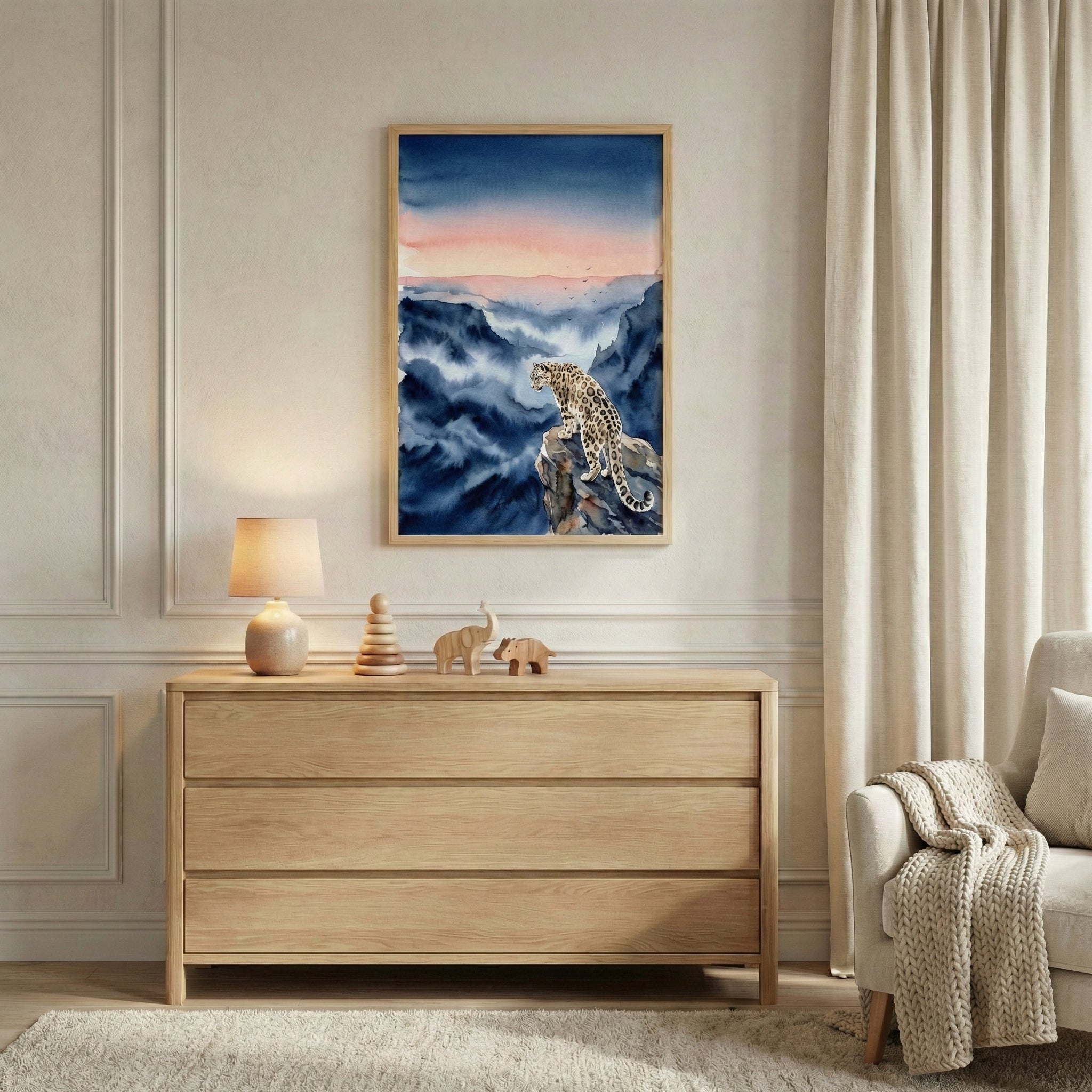 A bright, minimalist room features a light-colored wooden dresser against a paneled wall. On the dresser sit a small lamp, wooden animal toys, a stacking toy, and a stack of colorful children's books. Above the dresser hangs a framed watercolor-style artwork of a snow leopard perched on a rocky ledge, overlooking misty blue mountains under a sunset sky. To the right, long neutral curtains hang beside a plush armchair with a knitted throw. A soft light-colored rug rests on the floor.