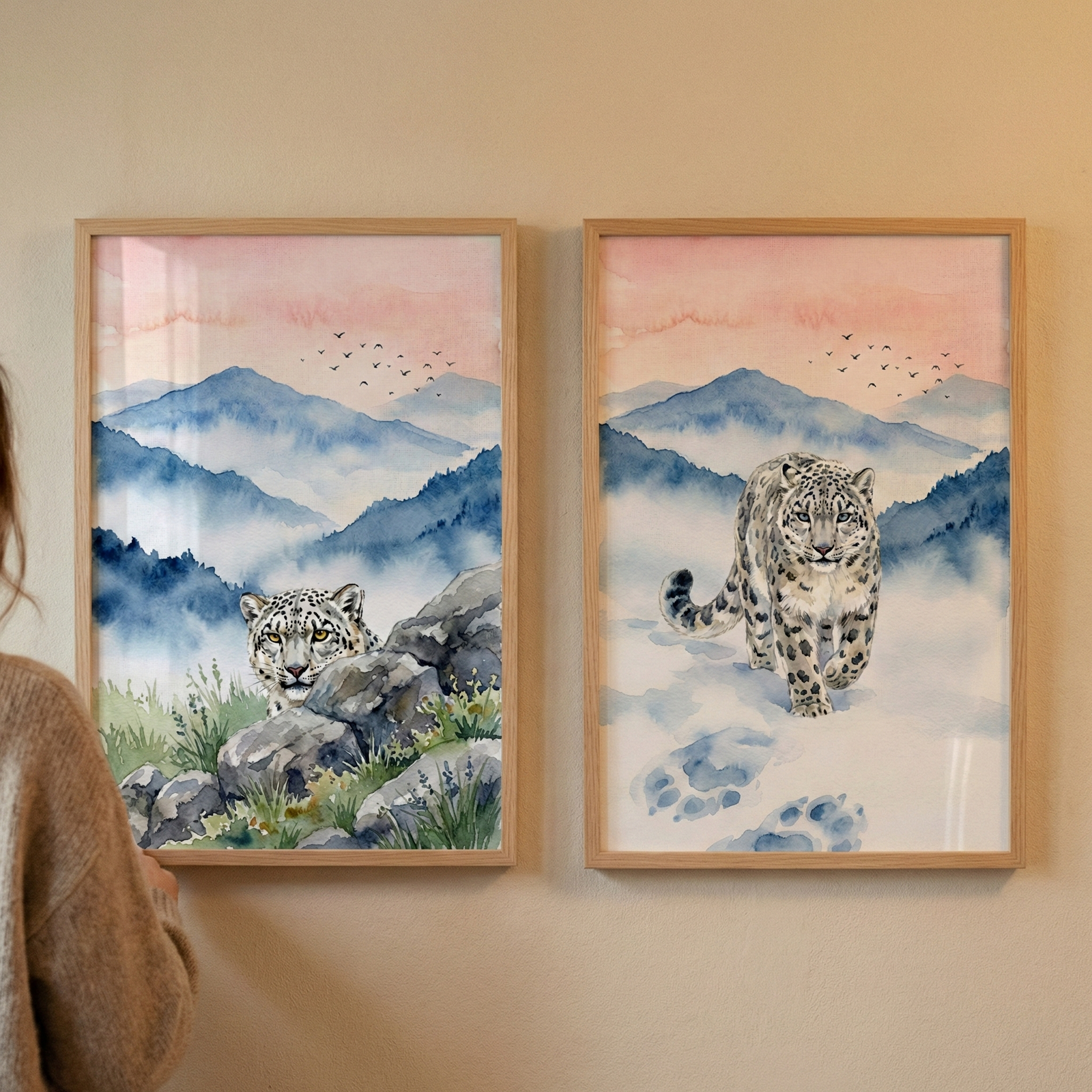 Two vertical, wood-framed watercolor illustrations hang side-by-side on a light wall. Both depict a serene mountain scene under a soft pink sky with small birds in flight. The left artwork shows a snow leopard peeking out from behind grey rocks and green grass. The right artwork features a snow leopard walking forward across a snowy landscape, leaving large blue paw prints behind it. A person's shoulder and arm are partially visible on the far left, as they reach towards the frames.