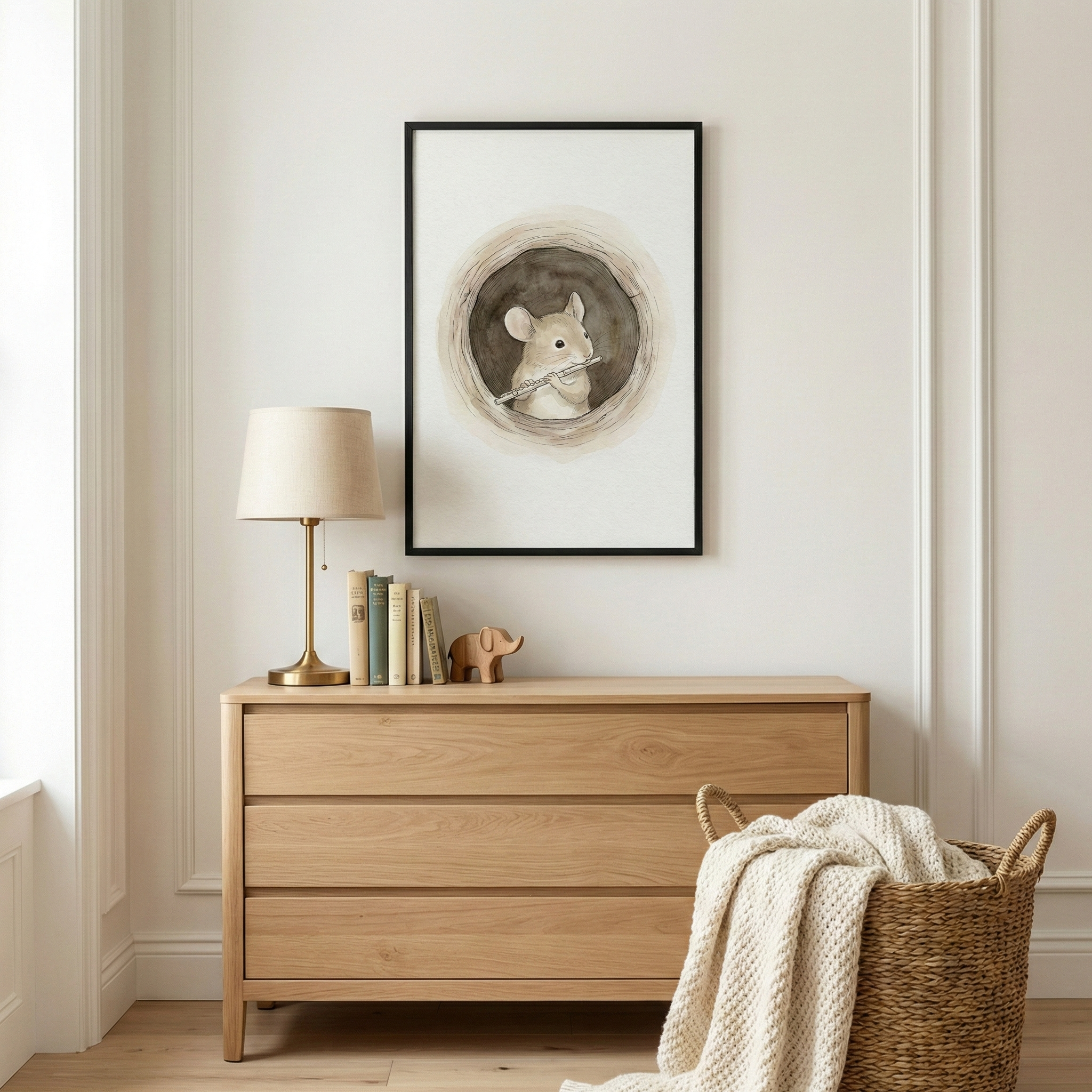 A well-lit, minimalist nursery corner features a three-drawer light-wood dresser against a neutral wall with molding. On the dresser sits a golden lamp with a beige shade, several books, and a small wooden elephant figurine. Hanging above is a black-framed illustration of a small, round-eared mouse inside a circular burrow, holding a thin flute. To the right of the dresser, a large woven basket holds a cream-colored, chunky knit blanket. The scene is set on a light wood floor.