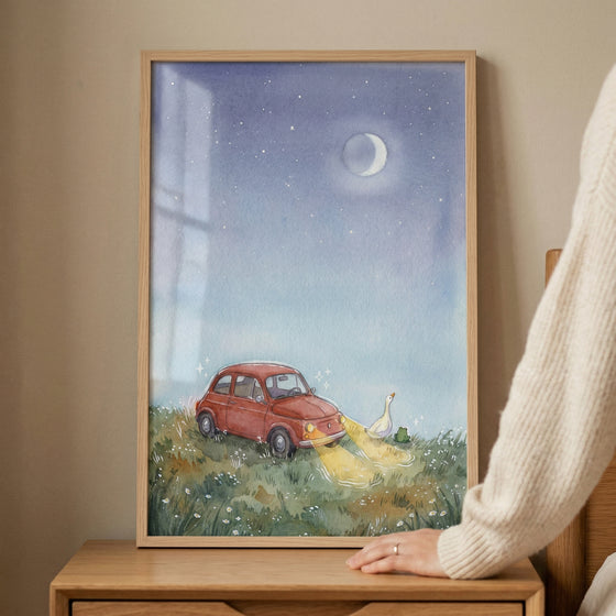 A framed watercolor-style illustration is displayed on a light wooden table. Inside the frame, a small red vintage car is depicted parked on a grassy field under a starry night sky. The car's headlights are turned on, casting a soft yellow glow onto the grass. In the path of the light, a white goose and a tiny green frog are visible. 