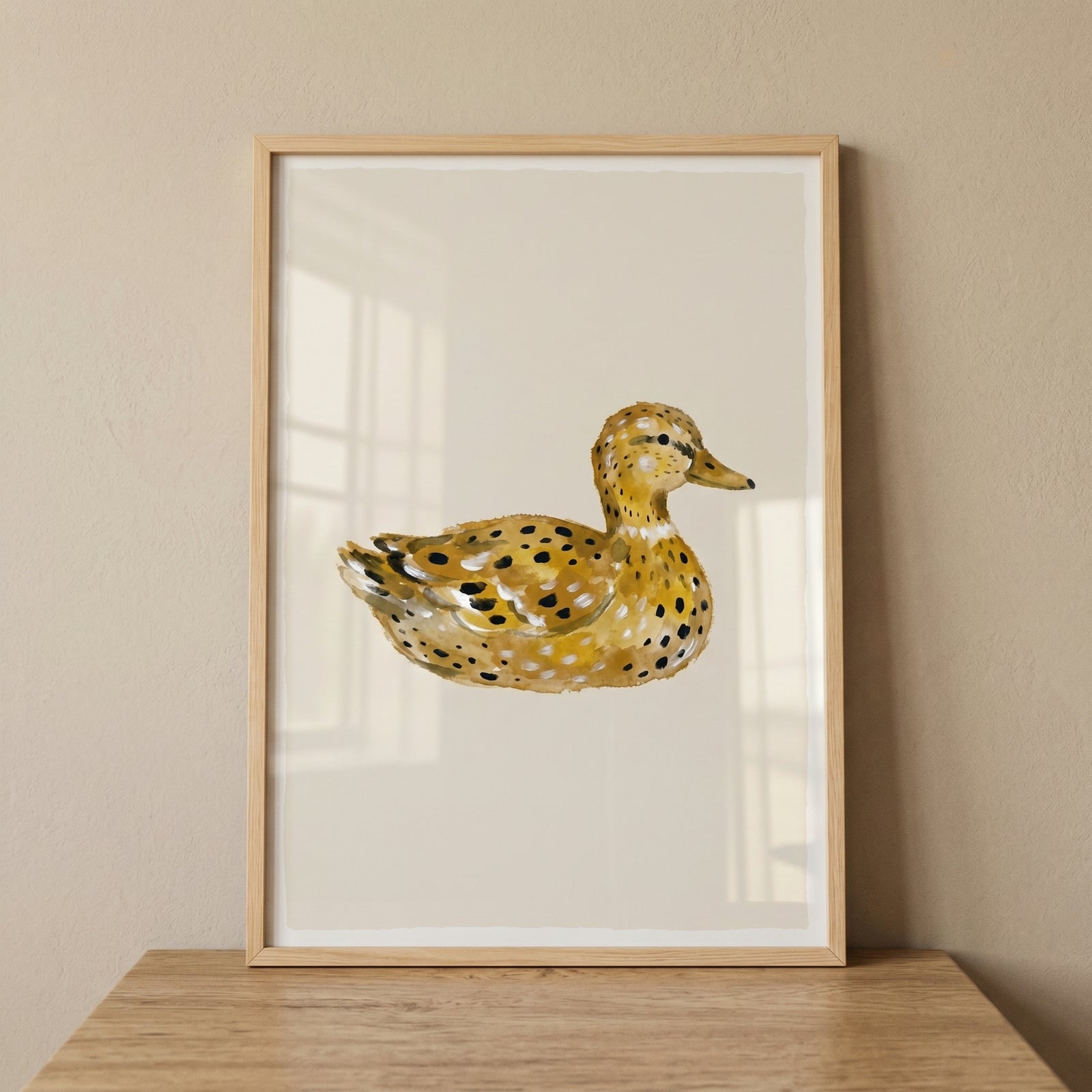 Framed watercolor ochre spotted duck wall art displayed on a wood ledge against a neutral backdrop.