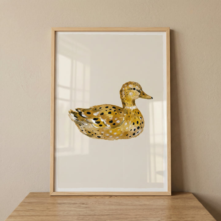 Framed watercolor ochre spotted duck wall art displayed on a wood ledge against a neutral backdrop.