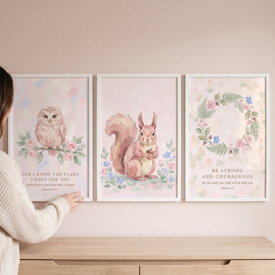 Hand hanging a watercolor petal forest nature artwork set of 3 featuring an owl, squirrel, and floral wreath.