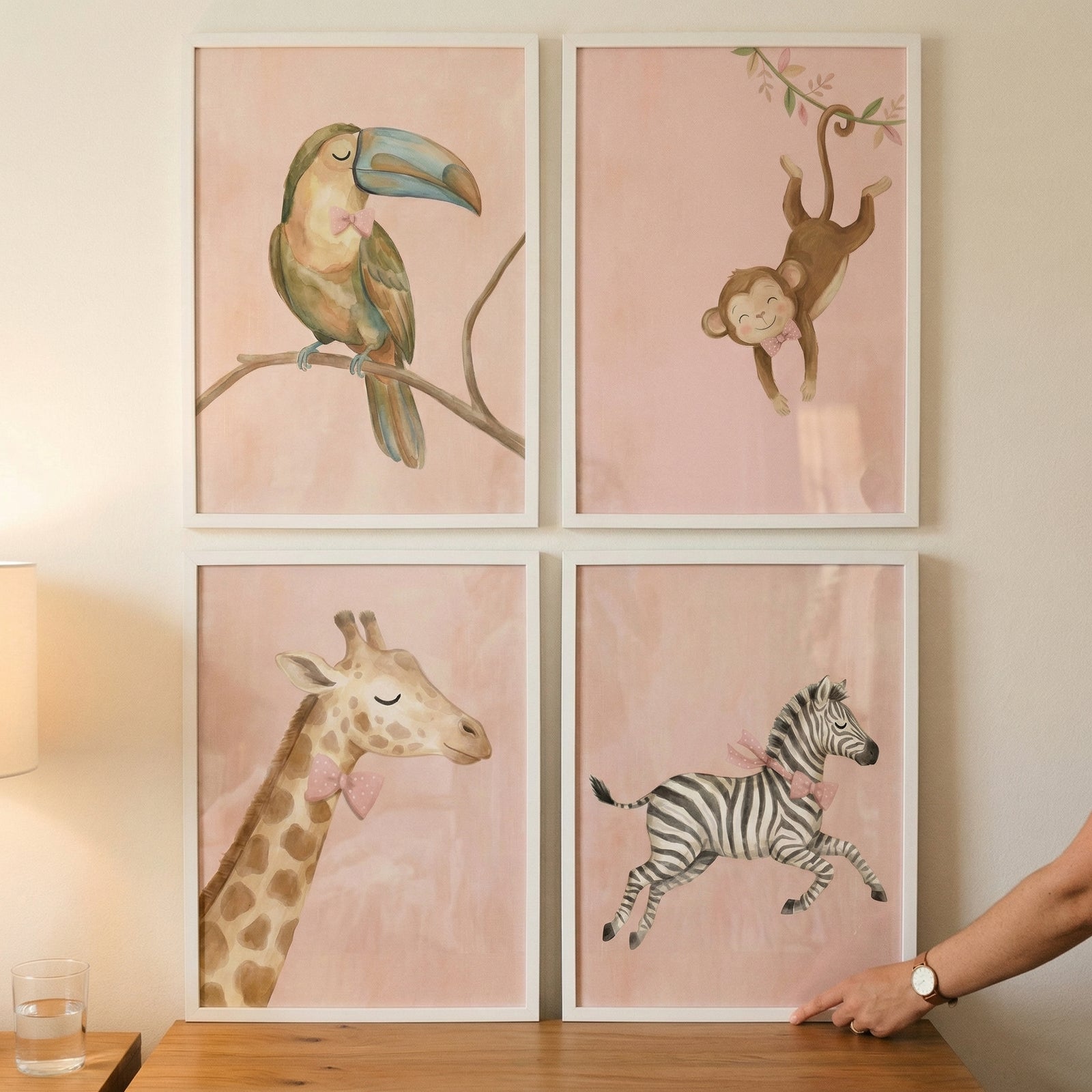 A set of four framed watercolor-style animal illustrations is displayed in a two-by-two grid on a neutral-toned wall. Each piece of art features a cute, whimsical animal depicted with soft colors against a textured light pink background.