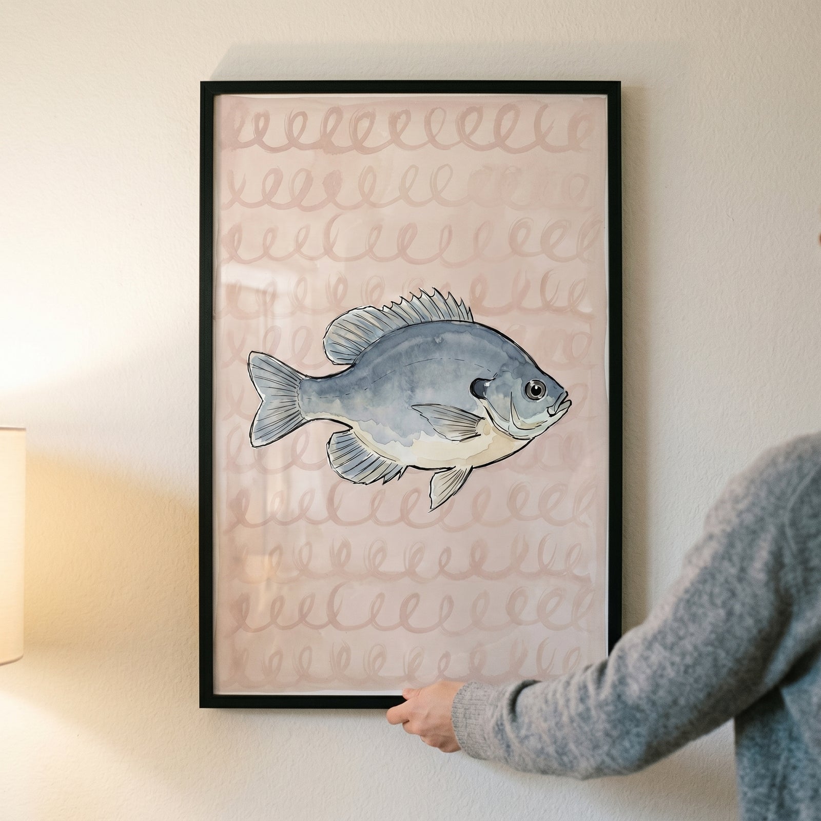 Hands holding watercolor pink spiral bluegill fish wall art in a black frame against a neutral indoor wall.