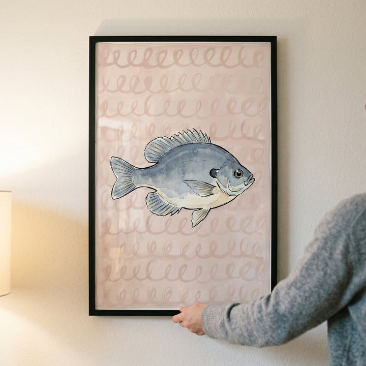 Hands holding watercolor pink spiral bluegill fish wall art in a black frame against a neutral indoor wall.
