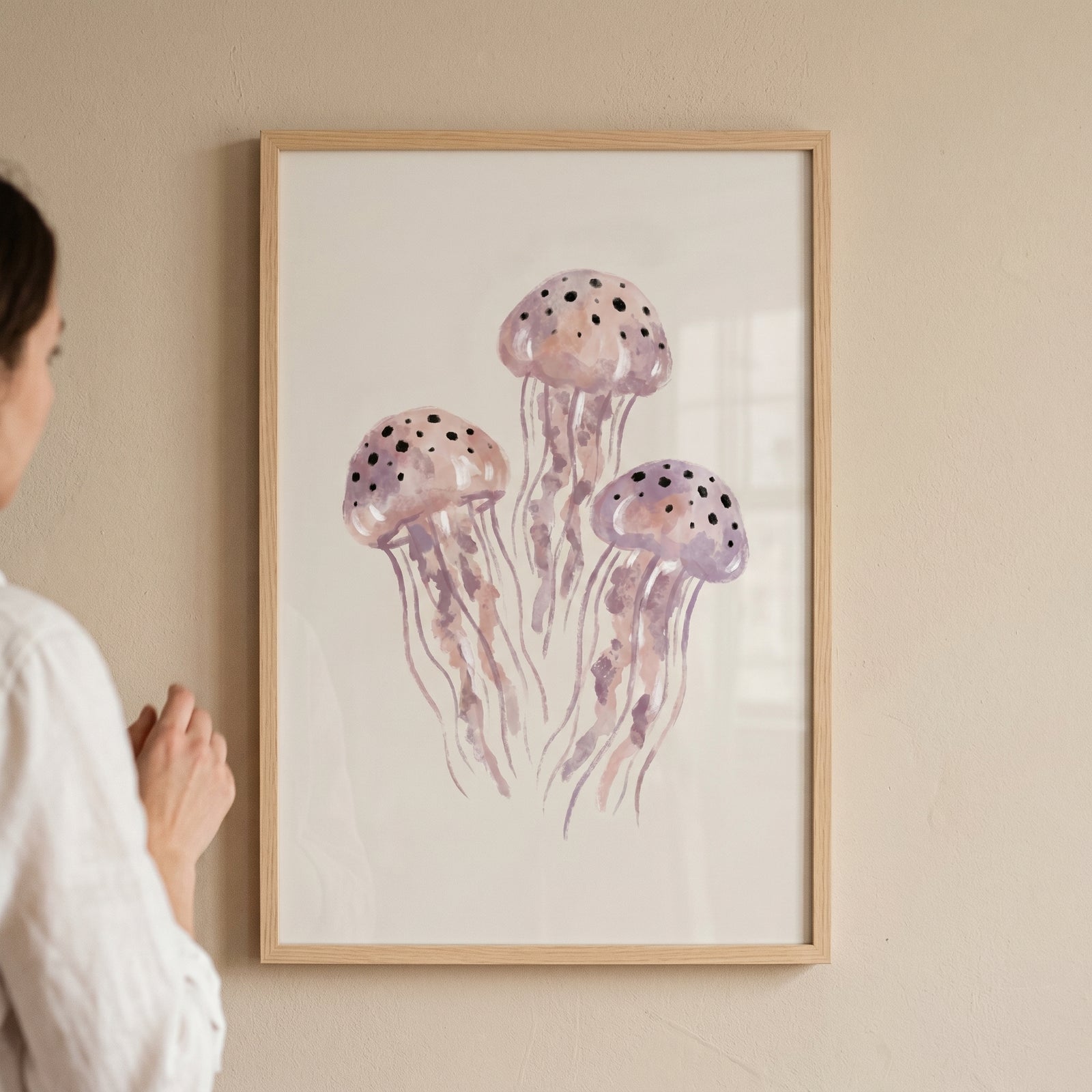 Watercolor purple jellyfish wall art in a light wood frame hangs on a beige wall next to a person.
