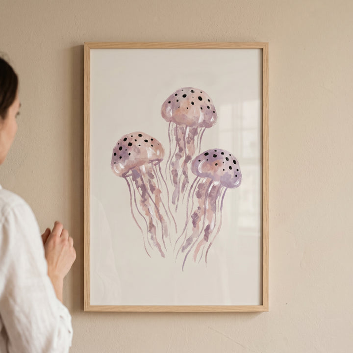Watercolor purple jellyfish wall art in a light wood frame hangs on a beige wall next to a person.