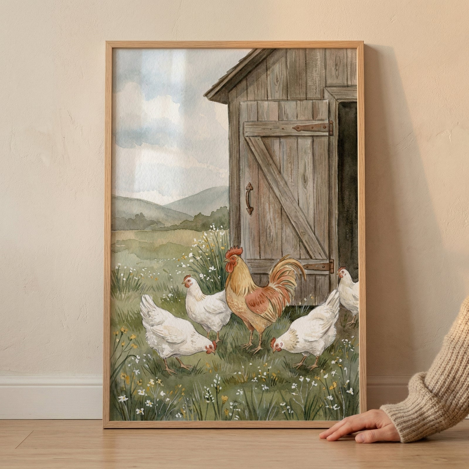 A framed watercolor illustration depicts a serene countryside scene. In the foreground, a vibrant rooster with golden-brown plumage and a large red comb stands prominently among four white hens. Some of the hens are pecking at the ground, foraging in a lush green field dotted with small white and yellow wildflowers.

