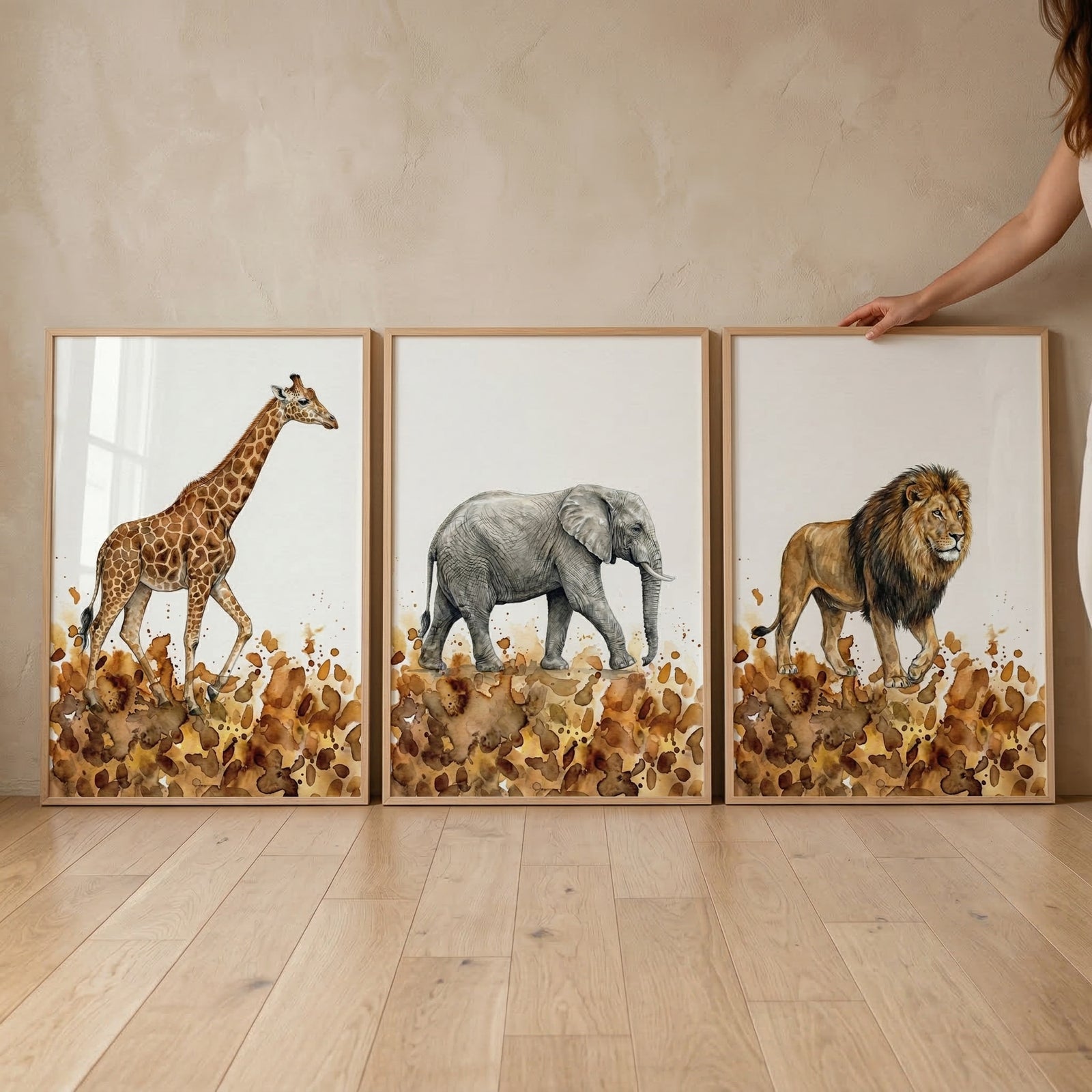 A set of three vertical framed illustrations leaning against a neutral, textured wall on a light wood floor. Each frame features a detailed, watercolor-style depiction of a safari animal on a clean white background. At the bottom of each artwork, there is an abstract ground composed of earthy brown and tan splotches. The left frame shows a giraffe walking to the right, the middle frame shows an elephant walking to the left, and the right frame shows a lion walking to the left. 