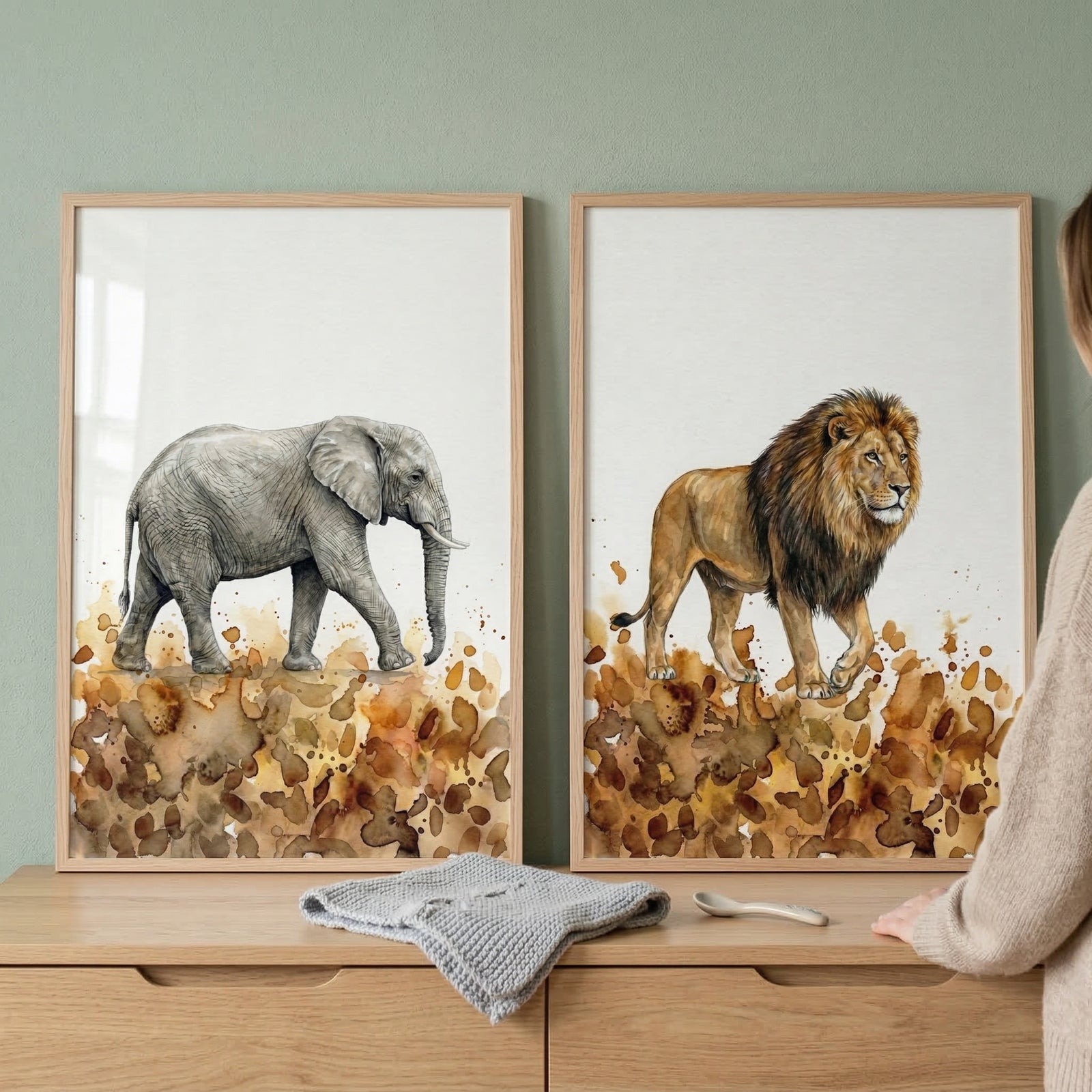 Two framed pieces of wall decor hang side-by-side on a soft green wall. The illustration on the left shows a gray elephant walking to the right, while the one on the right depicts a male lion with a dark mane walking to the left. Both animals are rendered in a watercolor style with abstract, earth-toned brown and tan splashes at their feet. The artworks are housed in light-colored wooden frames and positioned above a wooden chest of drawers.