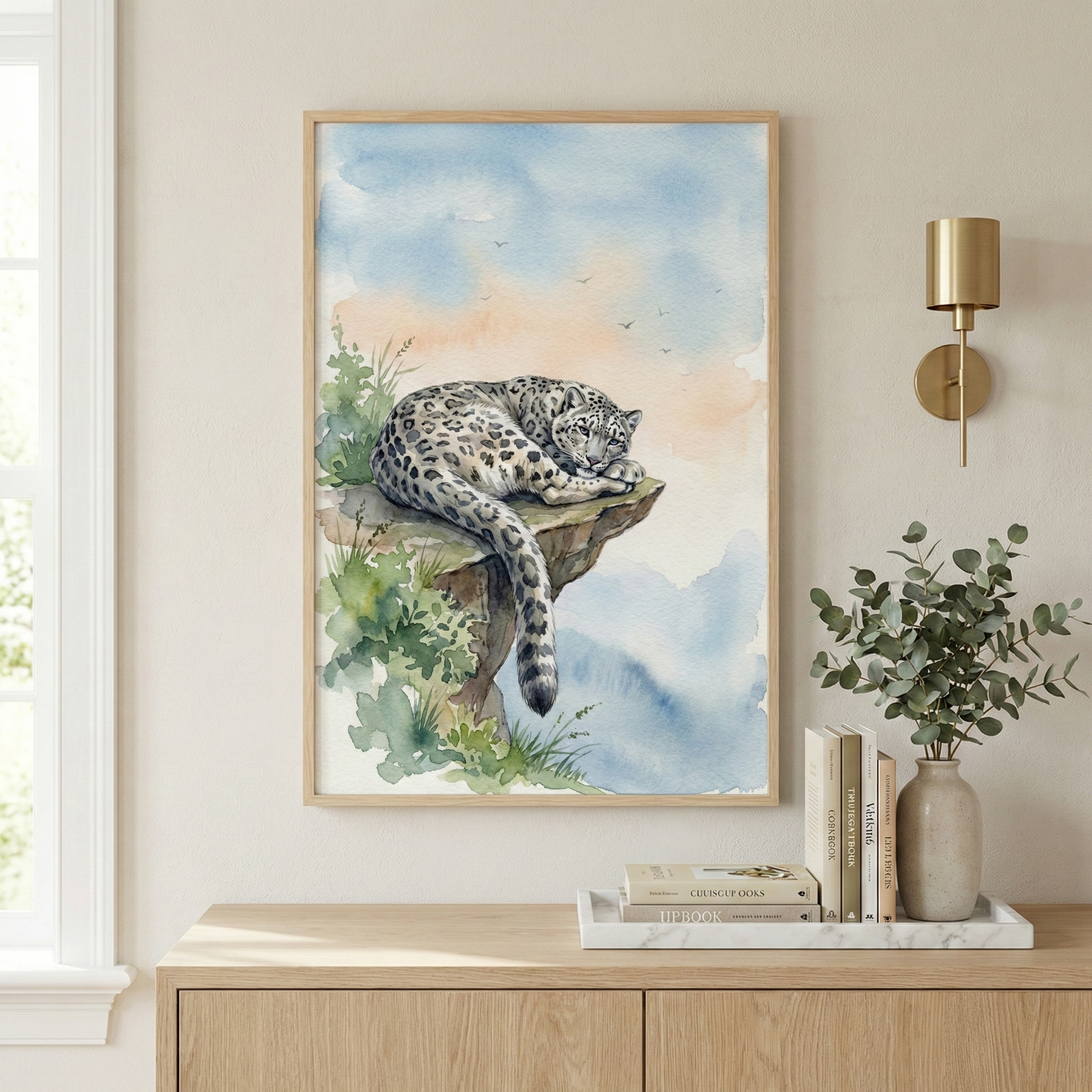 A framed watercolor painting of a snow leopard resting on a rocky ledge, surrounded by greenery, hangs on a light-colored wall. The background features a serene sky with soft blue and peach hues. A nearby wooden sideboard holds a vase with eucalyptus branches and a marble tray, under a brass wall sconce.