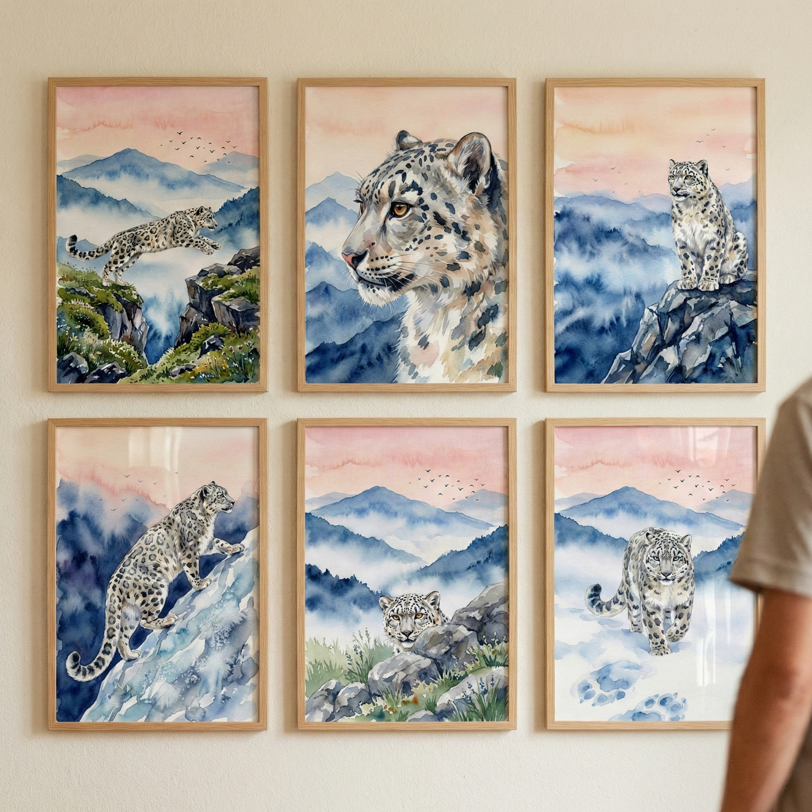 Six wooden-framed artworks are arranged in a 3x2 grid on a plain wall. Each watercolor-style illustration depicts a snow leopard in a misty mountain setting under a soft pink sky. The individual pieces show the leopard leaping across a gap, a close-up portrait, sitting on a peak, climbing a snowy slope, peeking from behind rocks, and walking forward in snow leaving paw prints. A person's shoulder is partially visible on the far right.