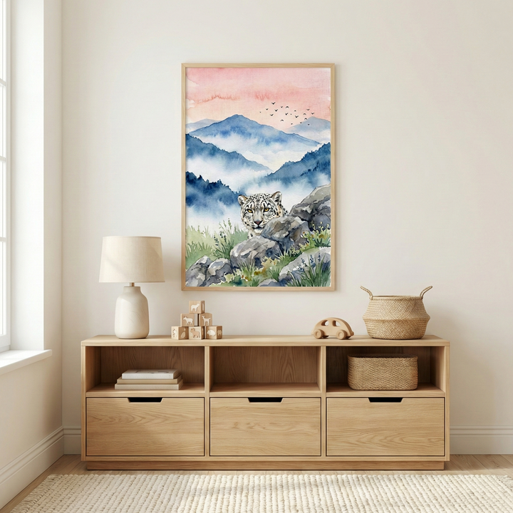 A framed watercolor-style illustration of a snow leopard hangs on a light-colored wall above a wooden sideboard. The leopard is shown peering over gray rocks in a mountain landscape with misty blue peaks and a soft pink and orange sky. The sideboard features three open compartments containing books, wooden blocks, and a woven basket, with three drawers underneath. A lamp with a white base and neutral shade sits on the left, alongside small wooden toys. A woven basket sits on the right side of the sideboard.