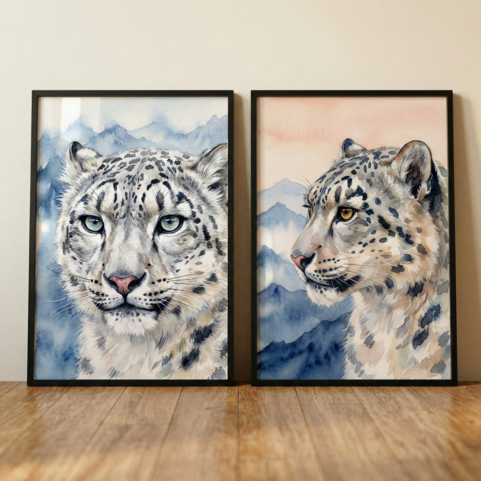 A pair of framed artworks featuring snow leopards in a watercolor style, leaning against a neutral wall on a light wood floor. The left illustration shows a front-facing snow leopard with pale eyes and blue mountain peaks in the background. The right illustration shows the animal in profile with amber eyes, set against blue mountains and a soft pink sky. Both pieces feature delicate spotted patterns and a soft, textured appearance.