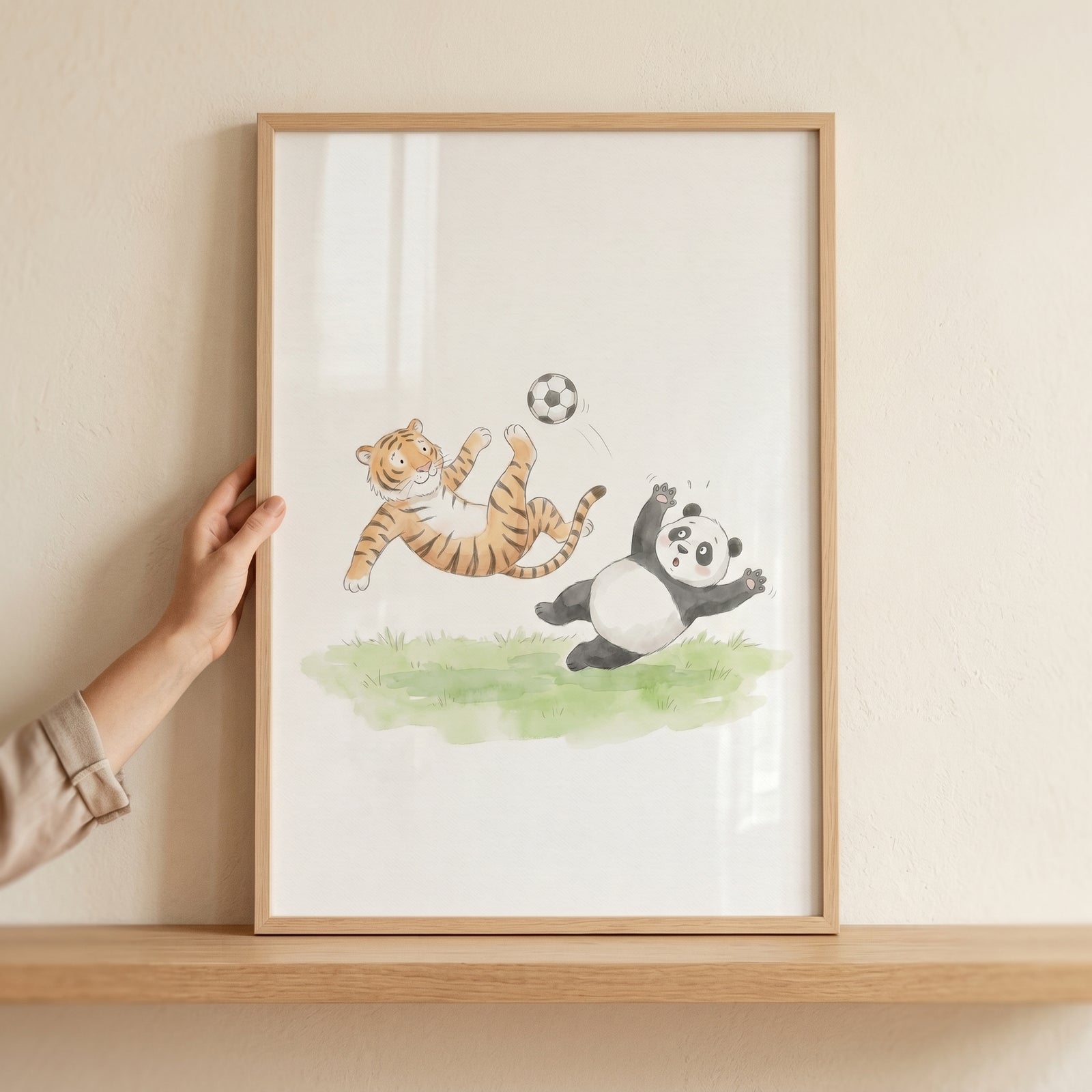 Watercolor soccer animals wall art depicts a tiger and panda playing soccer, in a wooden frame held by a hand.