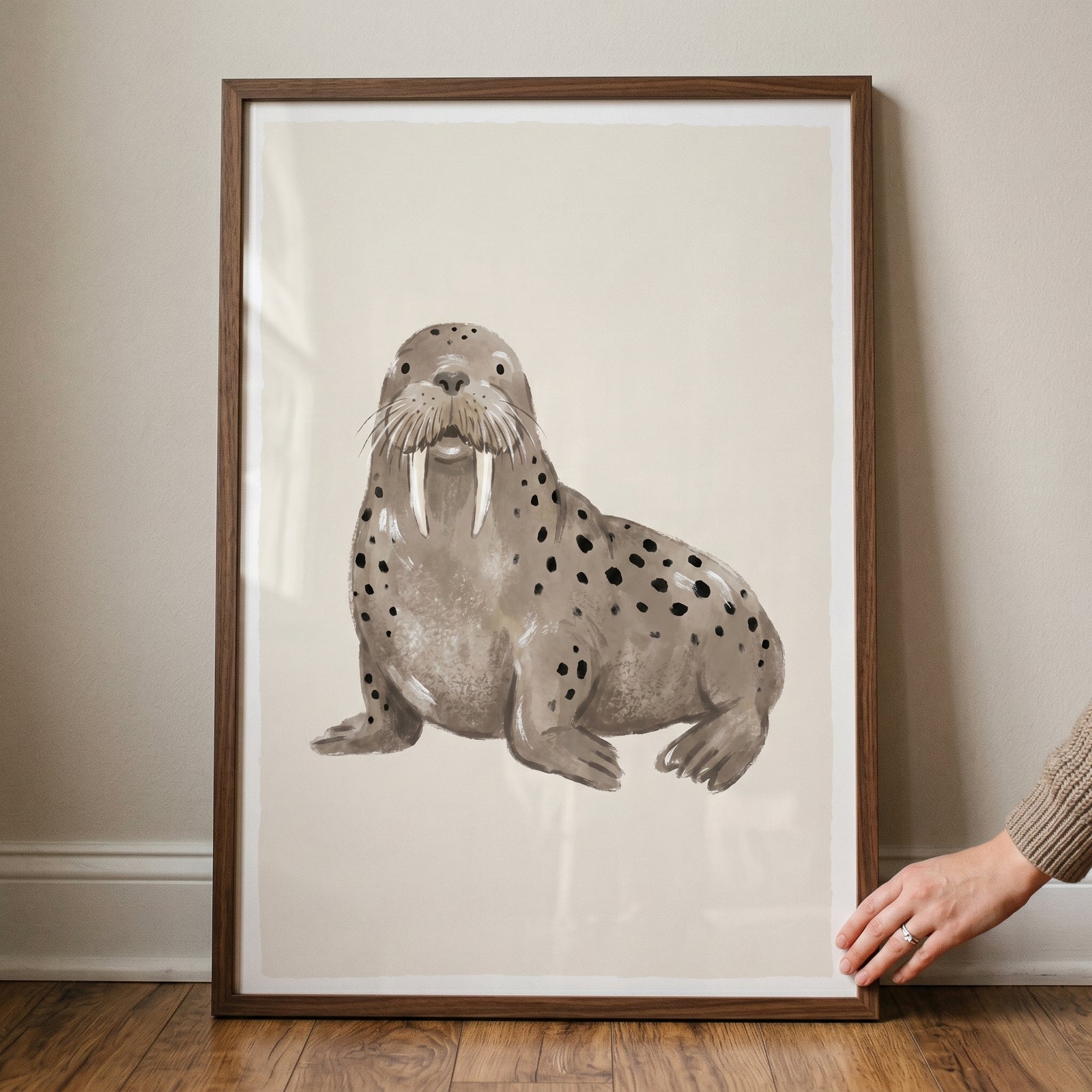 Watercolor spotted brown walrus wall art in a wooden frame resting on a wooden floor against a light wall.