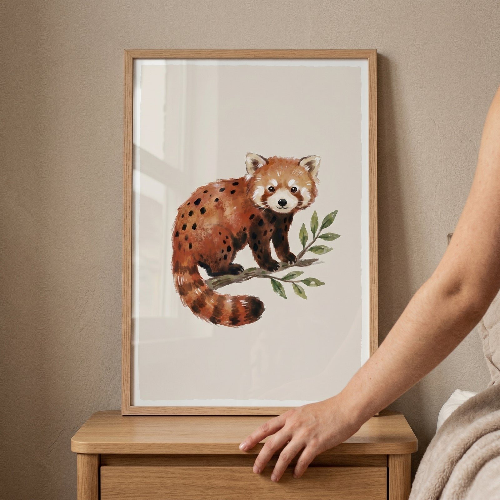 Watercolor spotted red panda wall art in a light wood frame resting on a wooden nightstand as a hand reaches out.
