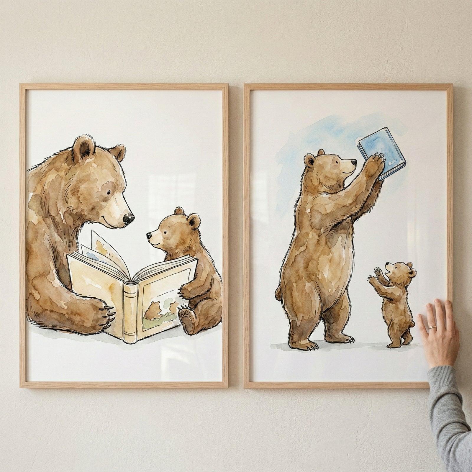 Two framed watercolor-style illustrations hang side-by-side on a light wall. In the left piece, a large brown bear and a small cub look at an open book together. The right piece shows the large bear standing on its hind legs, holding a blue book high above its head while the cub reaches up towards it. Both works feature soft textures and fine lines. The illustrations are in simple, light-colored wood frames. A hand is visible touching the edge of the right frame.
