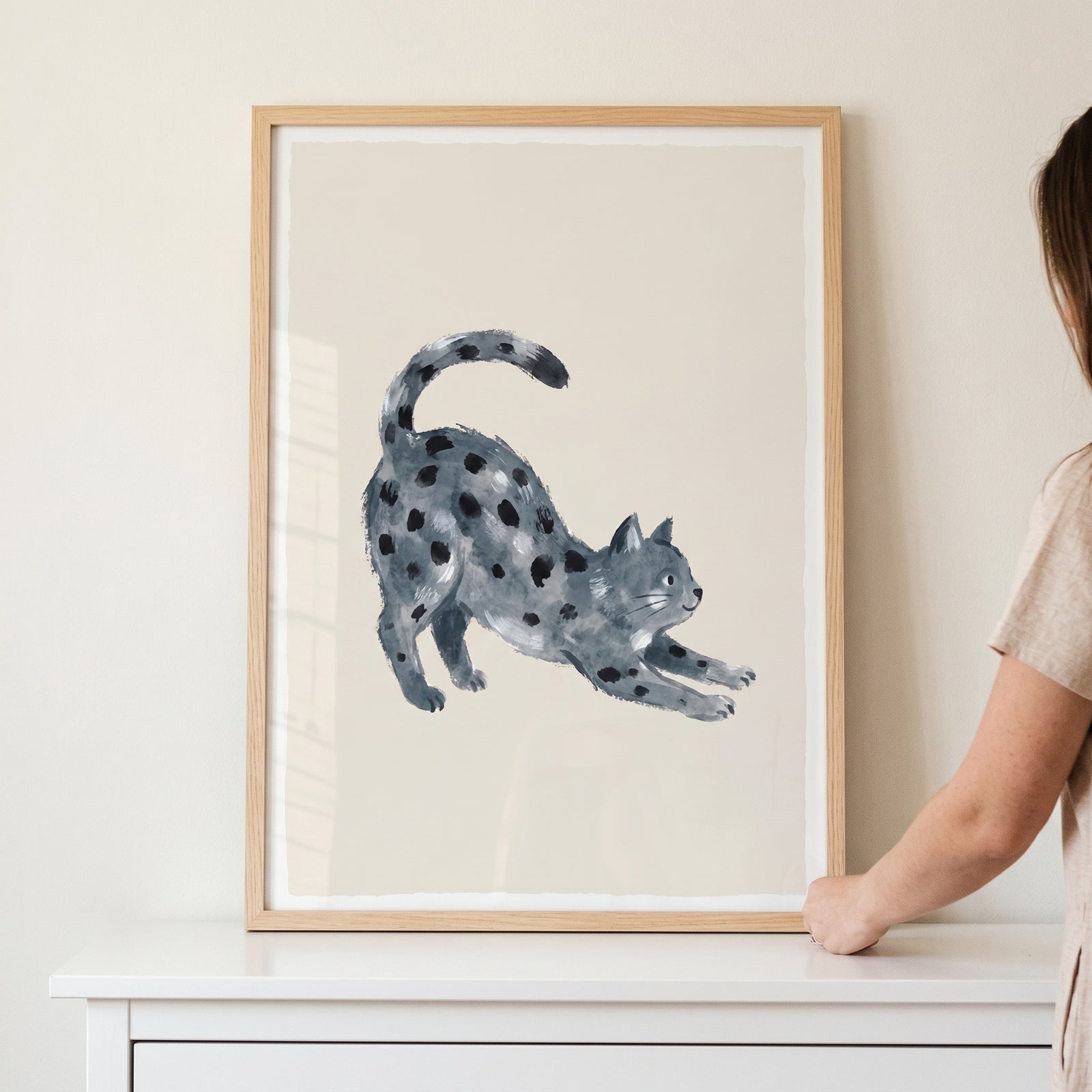 Framed watercolor stretching spotted cat wall art sits on a white dresser while a person holds the frame.