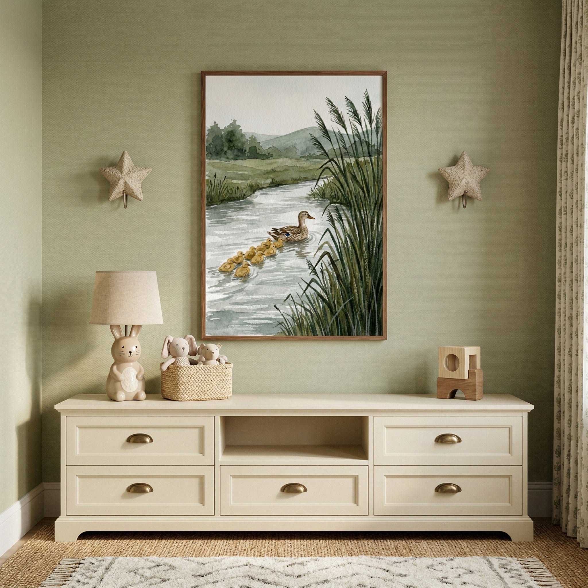 A serene, sage green nursery interior. Centered on the wall is a framed vertical illustration depicting a mother duck leading a line of yellow ducklings through a tranquil river, flanked by tall green reeds and distant soft hills. Two plush, star-shaped wall accents hang symmetrically on either side of the central frame. 
