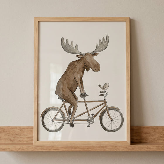 Framed watercolor tandem bike moose wall art shows a brown moose and a small bird on a bicycle, displayed on a wood shelf.