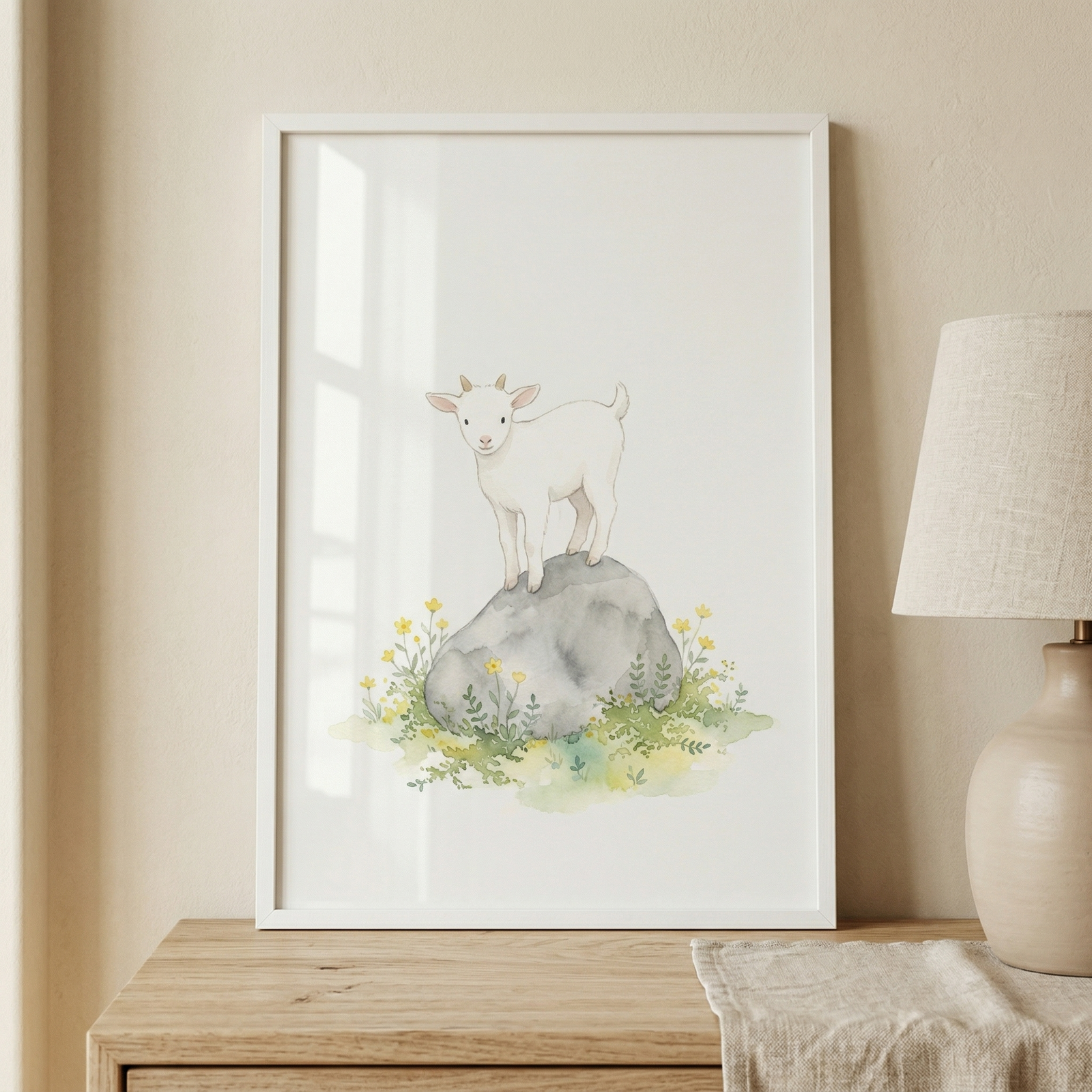 A framed watercolor illustration of a cute white baby goat standing atop a large grey rock. The rock sits amidst a small patch of grass with tiny yellow wildflowers. The illustration has a clean, minimalist aesthetic with a soft cream background. The thin white frame is resting on a light wood dresser against a neutral wall, positioned next to a modern lamp with a beige fabric shade and a round ceramic base. A piece of light linen fabric is draped on the dresser in front of the lamp.