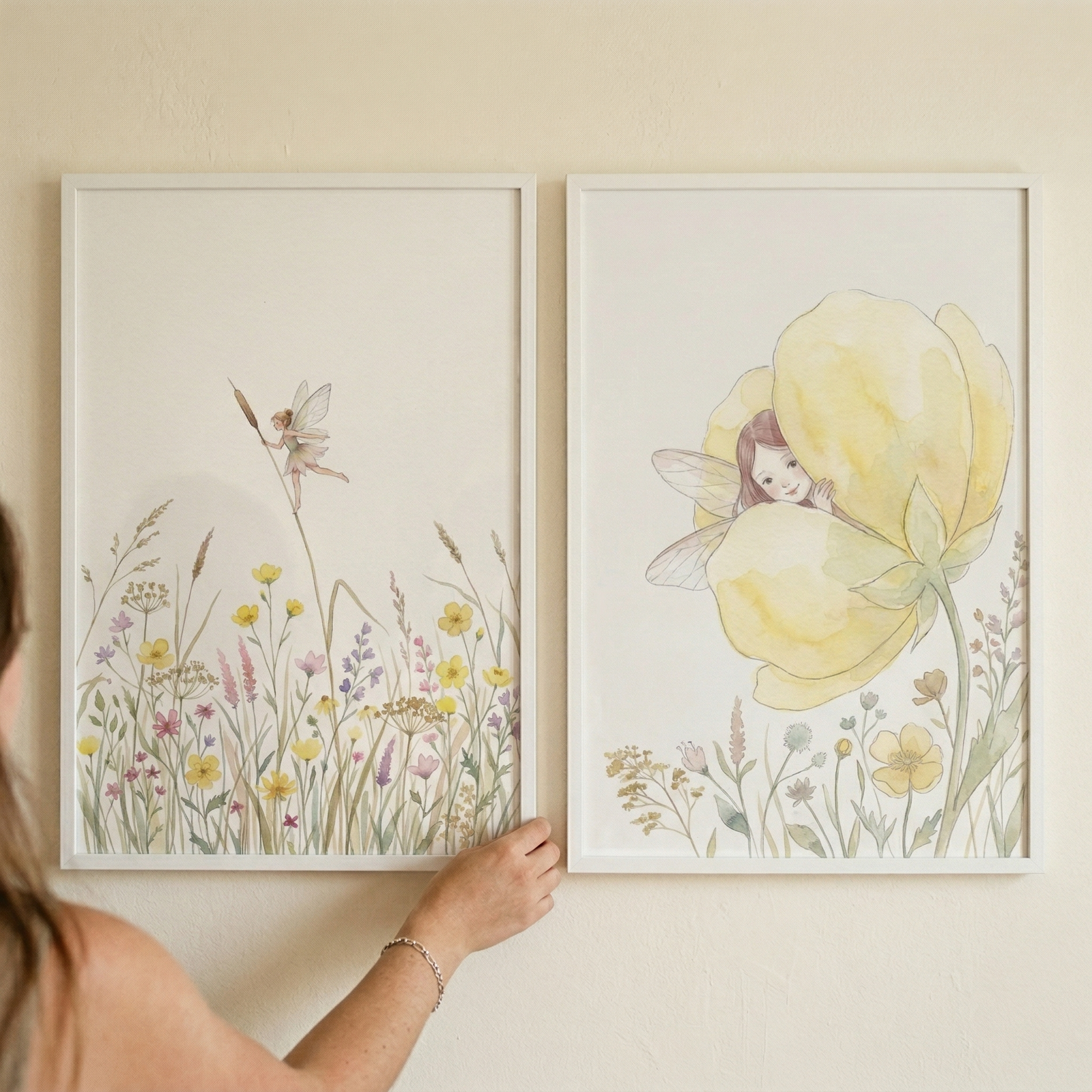 Two framed watercolor illustrations show delicate fairies in soft meadow scenes one flying above wildflowers while holding a cattail and the other peeking out from inside a large yellow flower. A hand adjusts one of the frames adding a styled real life touch to the gentle nursery decor setting.