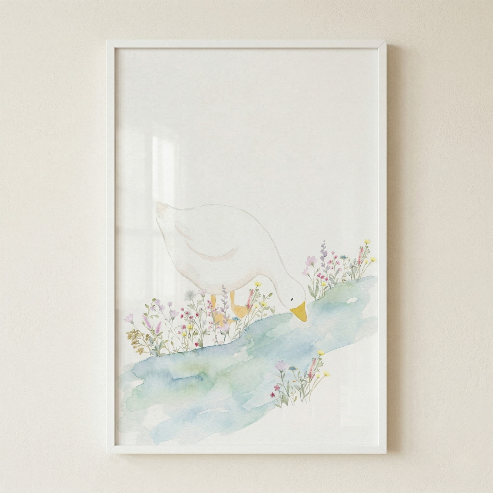 A framed watercolor illustration of a white duck leaning its head down toward a soft blue and green stream. Delicate wildflowers in shades of pink, yellow, and purple grow along the grassy bank where the duck stands. The artwork features a minimalist, airy style with a light background. It is housed in a simple white rectangular frame, displayed against a neutral, beige-colored wall.