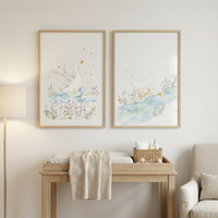 Two framed watercolor illustrations show white ducks in gentle nature scenes one splashing in water surrounded by small flowers and the other bending to drink from a stream. The prints hang above a light wood changing table with soft neutral decor creating a calm nature inspired nursery setting.