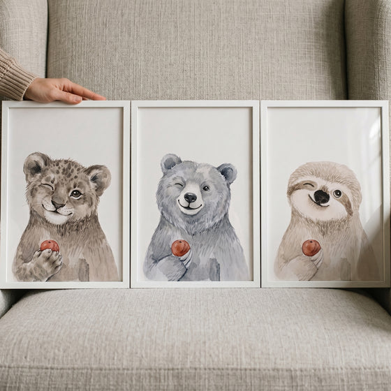 Watercolor winking baby animal wall art set of 3 featuring a leopard, bear, and sloth holding small red fruit.