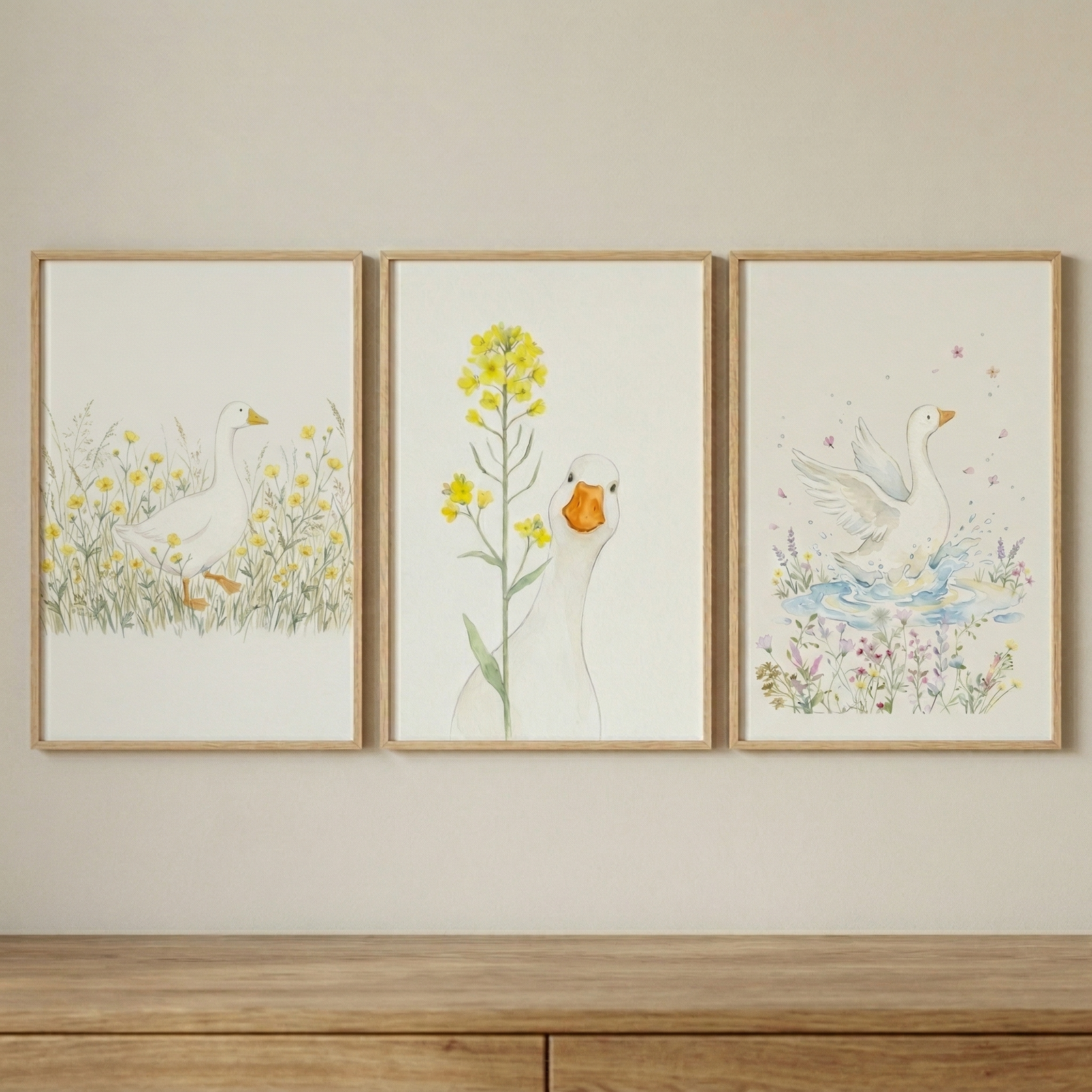 A set of three vertical, watercolor-style illustrations in light wood frames, hanging on a neutral-toned wall. Each art piece features a whimsical white duck in a different natural setting. The piece on the left shows a white duck strolling through a field of tall grass and yellow wildflowers. The central piece depicts a white duck peeking from behind a tall green plant with bright yellow flowers. The piece on the right illustrates a white duck with its wings outstretched, landing in a pool of water amidst 