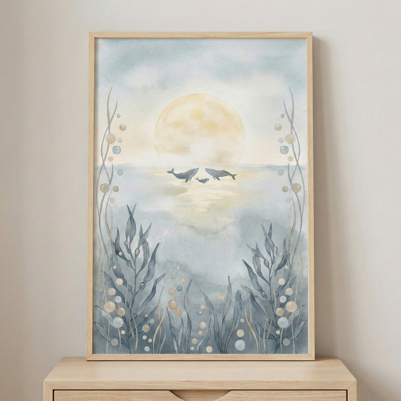 A vertical watercolor artwork is displayed in a light wood frame on a matching surface. The central focus is a large, soft yellow sun or moon low on the horizon, reflecting on calm water. Three whale silhouettes swim along the horizon line. The scene is framed by ethereal underwater foliage, including stalks of seaweed and floating bubbles in shades of blue and beige. The palette is soft and muted, creating a serene and whimsical atmosphere.