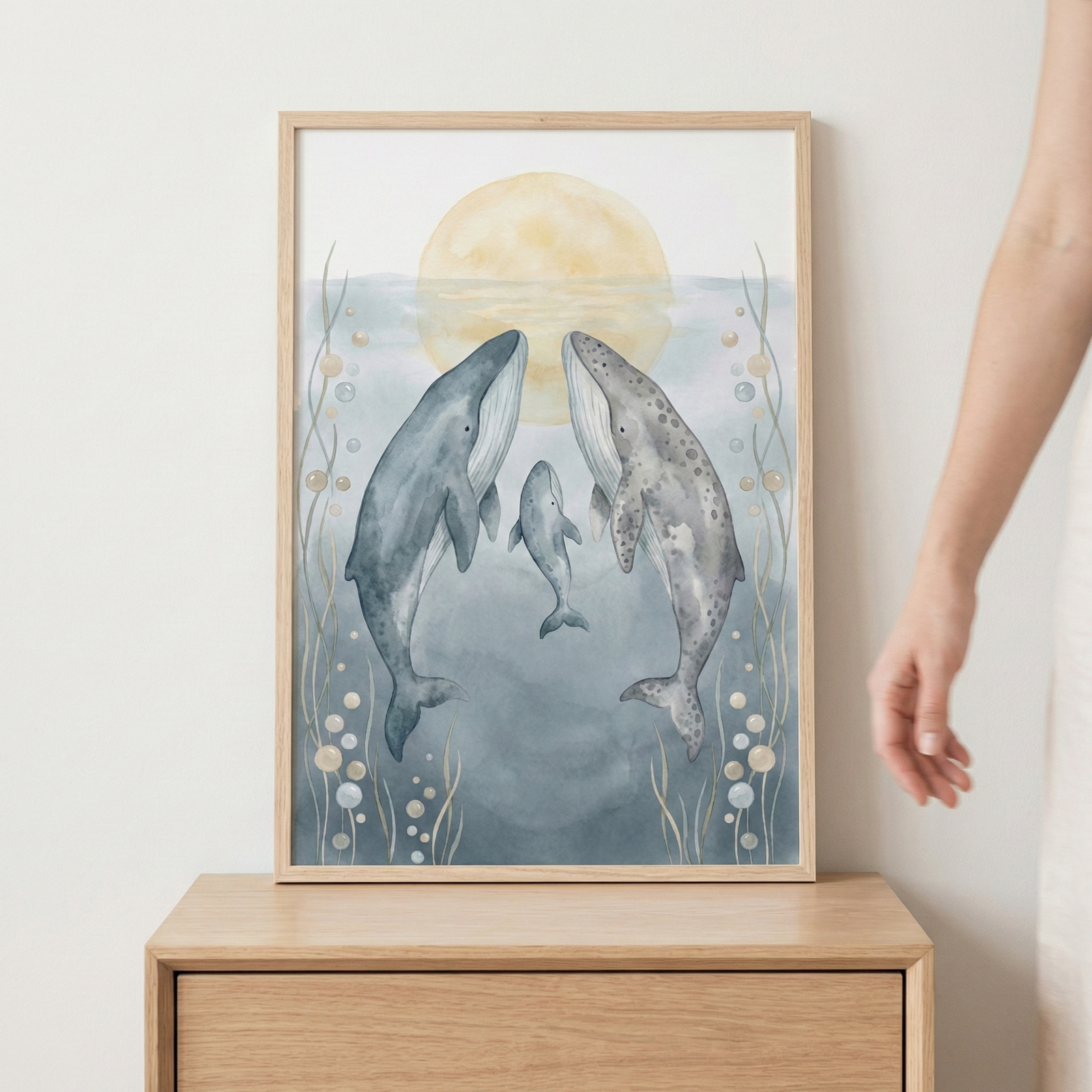 A framed watercolor-style illustration of a whale family consisting of two large whales and a small whale swimming together in a serene underwater setting. They are centered beneath a large, pale yellow sun or moon in a hazy blue-gray sea. The composition is decorated with delicate bubbles and slender aquatic plants on either side. The light-wood frame sits on a matching wooden dresser.