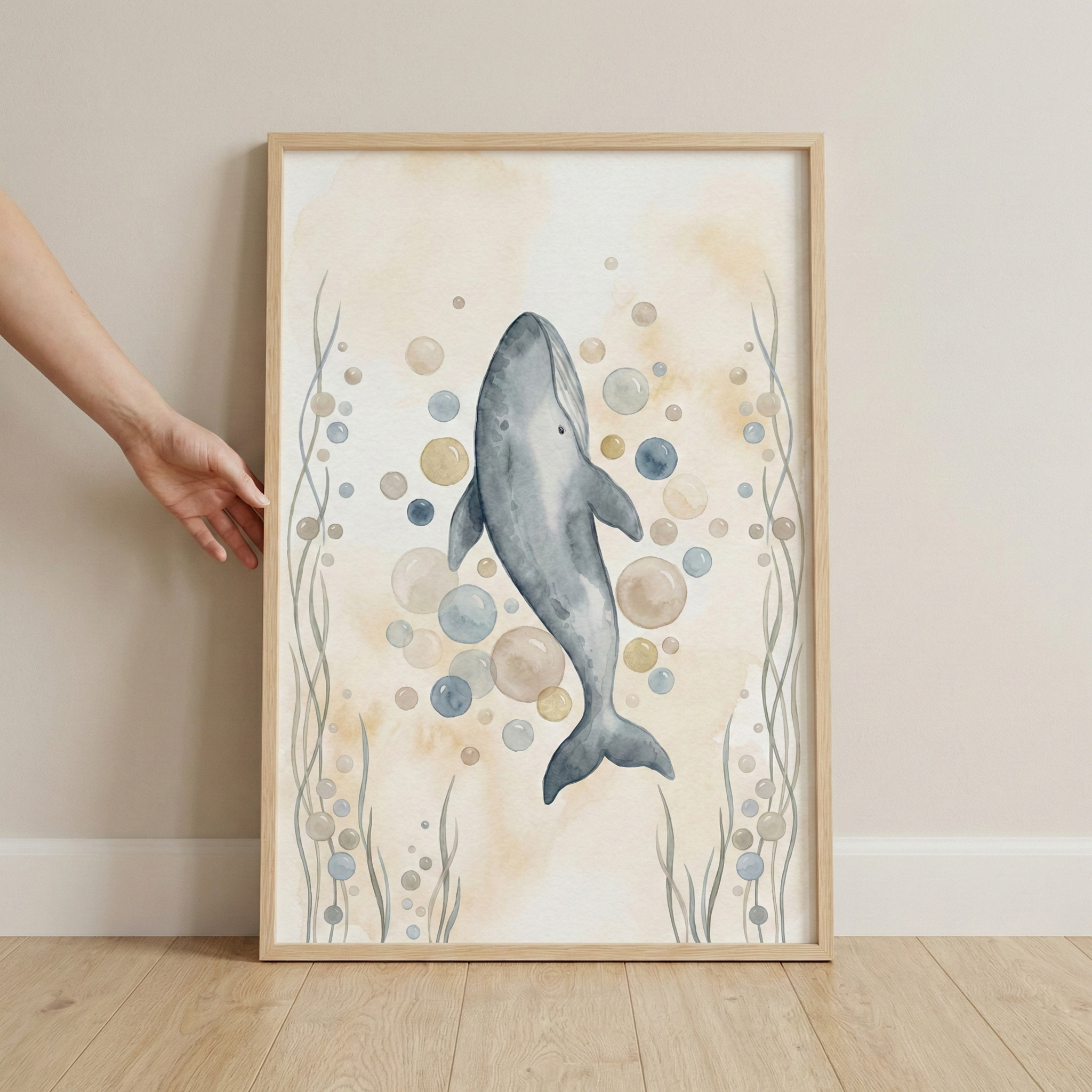 A vertical watercolor-style illustration of a large gray whale swimming upwards. The whale is surrounded by a cluster of bubbles in shades of blue, pale yellow, and beige. Long, wavy aquatic plants in gray and blue tones line the sides of the artwork. The piece is set within a light-colored wood frame, which is resting on a light wood floor against a neutral-colored wall. A person's hand and arm are visible on the left, touching the side of the frame.