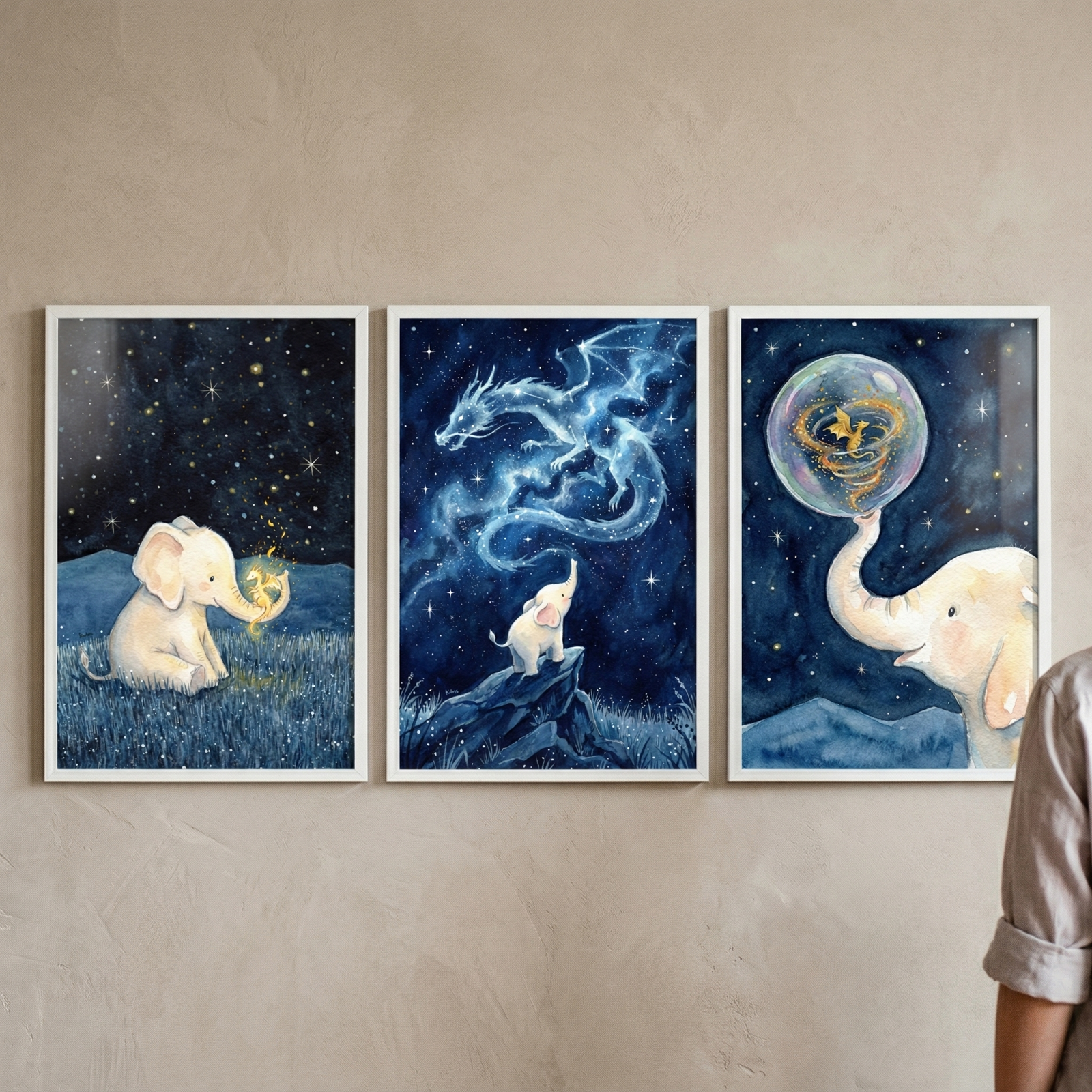 A triptych of three vertical white-framed illustrations hung side-by-side on a textured beige wall. The first piece on the left shows a small white elephant sitting in a field of dark blue grass under a night sky filled with twinkling stars, holding a tiny, glowing golden creature in its trunk. The center illustration depicts the small elephant standing on a rocky peak, gazing up at a majestic, ethereal dragon made of swirling blue stardust and light. 