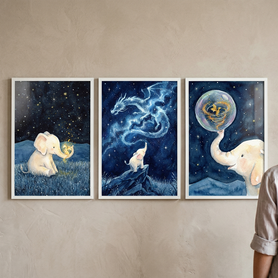 A triptych of three vertical white-framed illustrations hung side-by-side on a textured beige wall. The first piece on the left shows a small white elephant sitting in a field of dark blue grass under a night sky filled with twinkling stars, holding a tiny, glowing golden creature in its trunk. The center illustration depicts the small elephant standing on a rocky peak, gazing up at a majestic, ethereal dragon made of swirling blue stardust and light. 