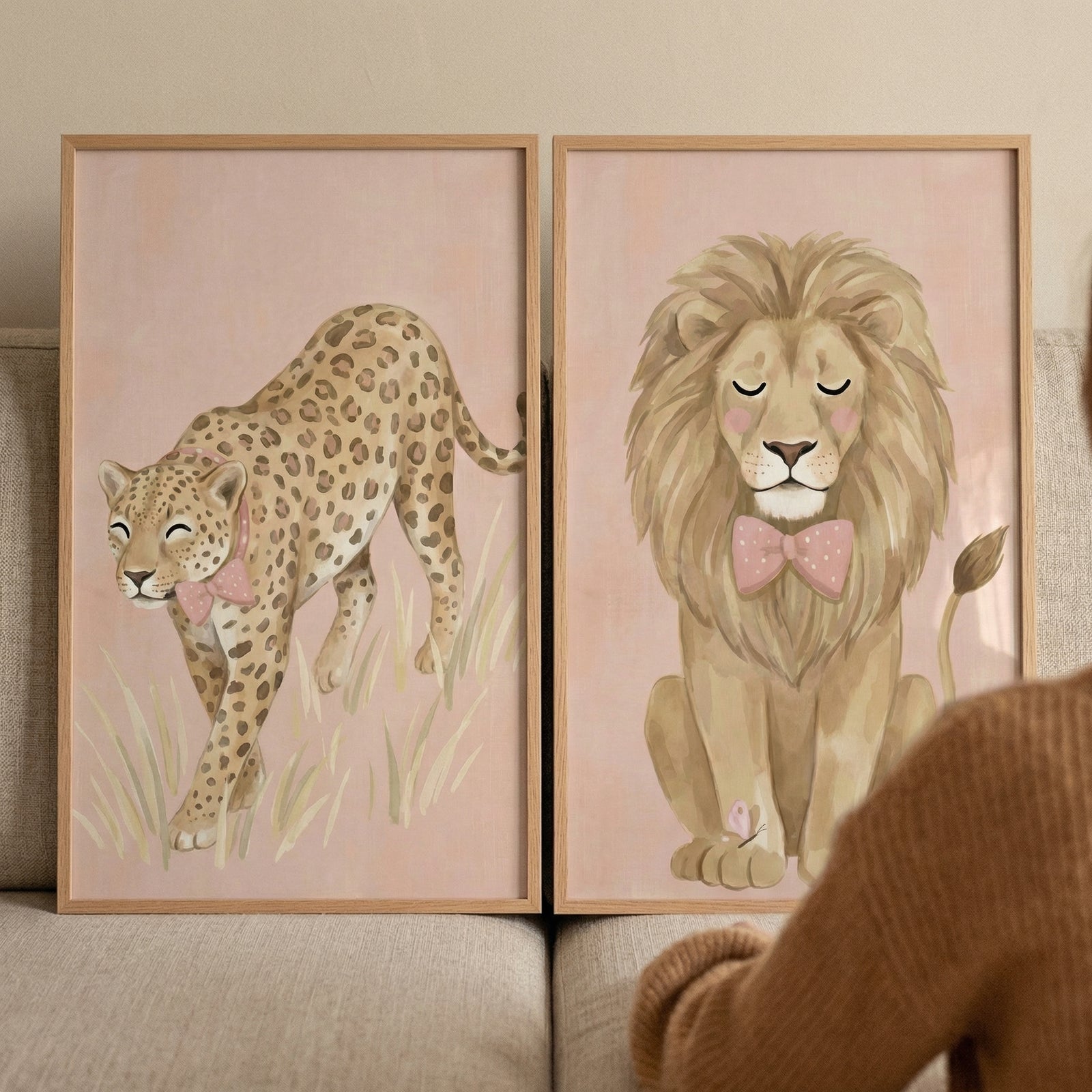 This pair of framed illustrations features whimsical and gentle depictions of large cats, perfect for a nursery or child's room. On the left, a soft-toned leopard with classic spots is depicted walking through a patch of tall, pale grass. It wears a charming pink bow around its neck, and the entire scene is set against a muted pink background.