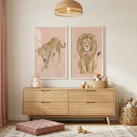A softly lit nursery features warm wood furniture and gentle pink tones. Two white-framed illustrations are mounted on the wall; the first depicts a leopard walking through grass, and the second shows a seated lion with a pink bow tie, both set against muted pink backgrounds.