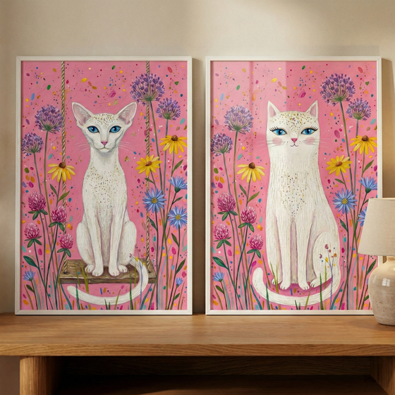 Two framed pieces of art displayed on a wooden surface show stylized white cats against bright pink backgrounds. Both cats have blue eyes and gold-speckled fur, surrounded by colorful wildflowers in shades of purple, yellow, and blue. On the left, a cat sits on a rustic wooden swing. On the right, a cat with long eyelashes sits with its tail curled forward. The background features playful, multicolored speckles. A lamp base is partially visible on the right.