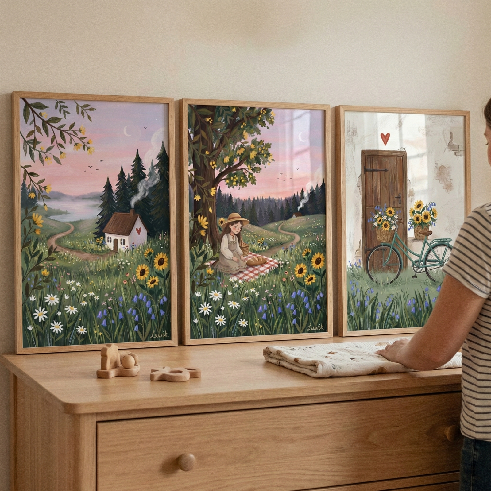 Three framed whimsical illustrations hang above a light wood dresser. The left frame shows a cottage in a flowery field under a pink sky. The middle one depicts a girl on a picnic blanket in a meadow under a tree. The right frame features a vintage-style blue bicycle with sunflowers in front of a wooden door. A person is partially visible, arranging a cloth on the dresser where small wooden toys are placed. The scene is calm and cozy with soft colors.