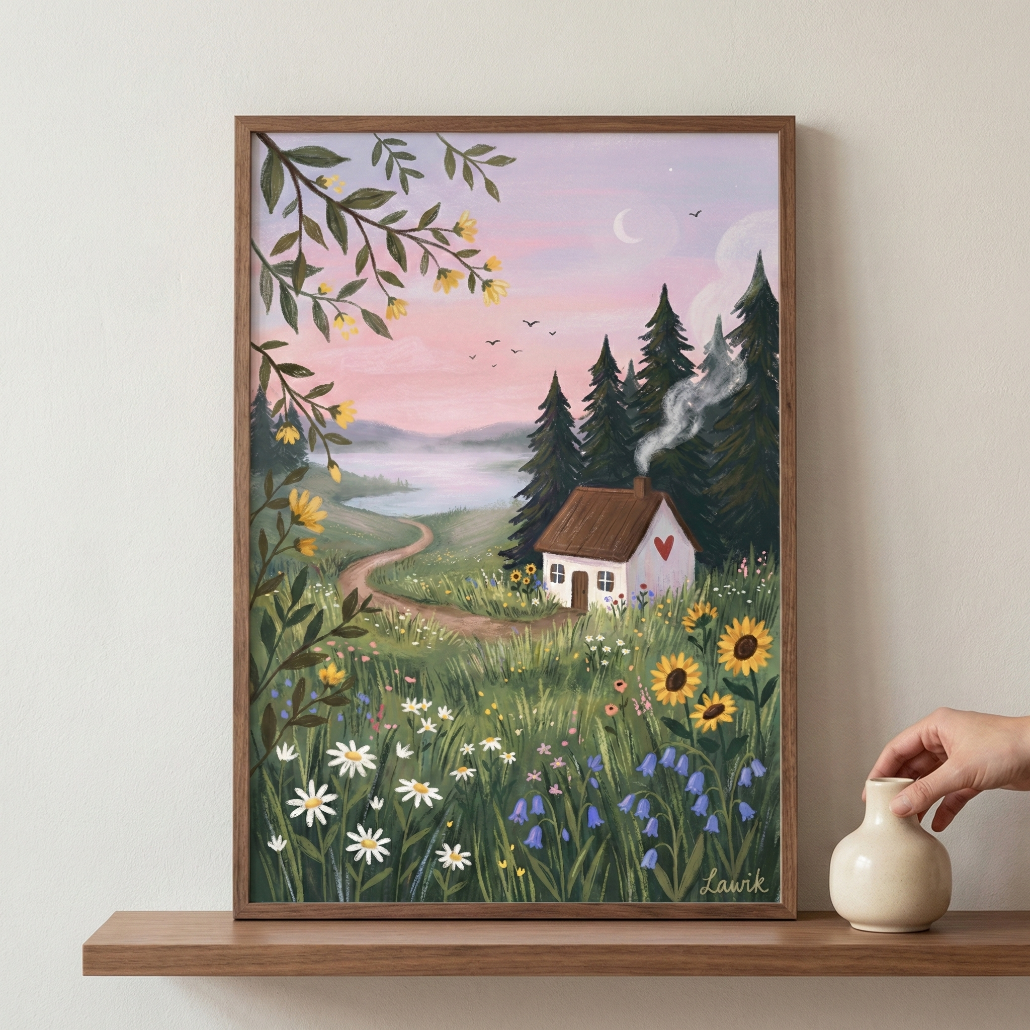 A framed illustration sits on a wooden shelf, depicting a whimsical countryside scene. A small white cottage with a red heart on its exterior is nestled in a vibrant meadow of sunflowers, daisies, and bluebells. A winding dirt path leads from the house toward a misty lake under a soft pink and purple sky featuring a slender crescent moon. Tall, dark evergreen trees and a thin wisp of chimney smoke enhance the cozy atmosphere. To the right, a hand reaches for a small white ceramic vase on the shelf.