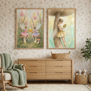 Two framed whimsical illustrations hang on a wall with a delicate floral pattern. The first illustration shows two sprites with delicate wings sitting on toadstools amidst tall grass, sharing a glowing orb. The second features a sprite leaning against a large toadstool stem while reading a book. Below the art is a light wood dresser with a wicker basket. To the left is a rocking chair with a green knit throw, and to the right is a potted plant and a basket of plush toys on a white rug.