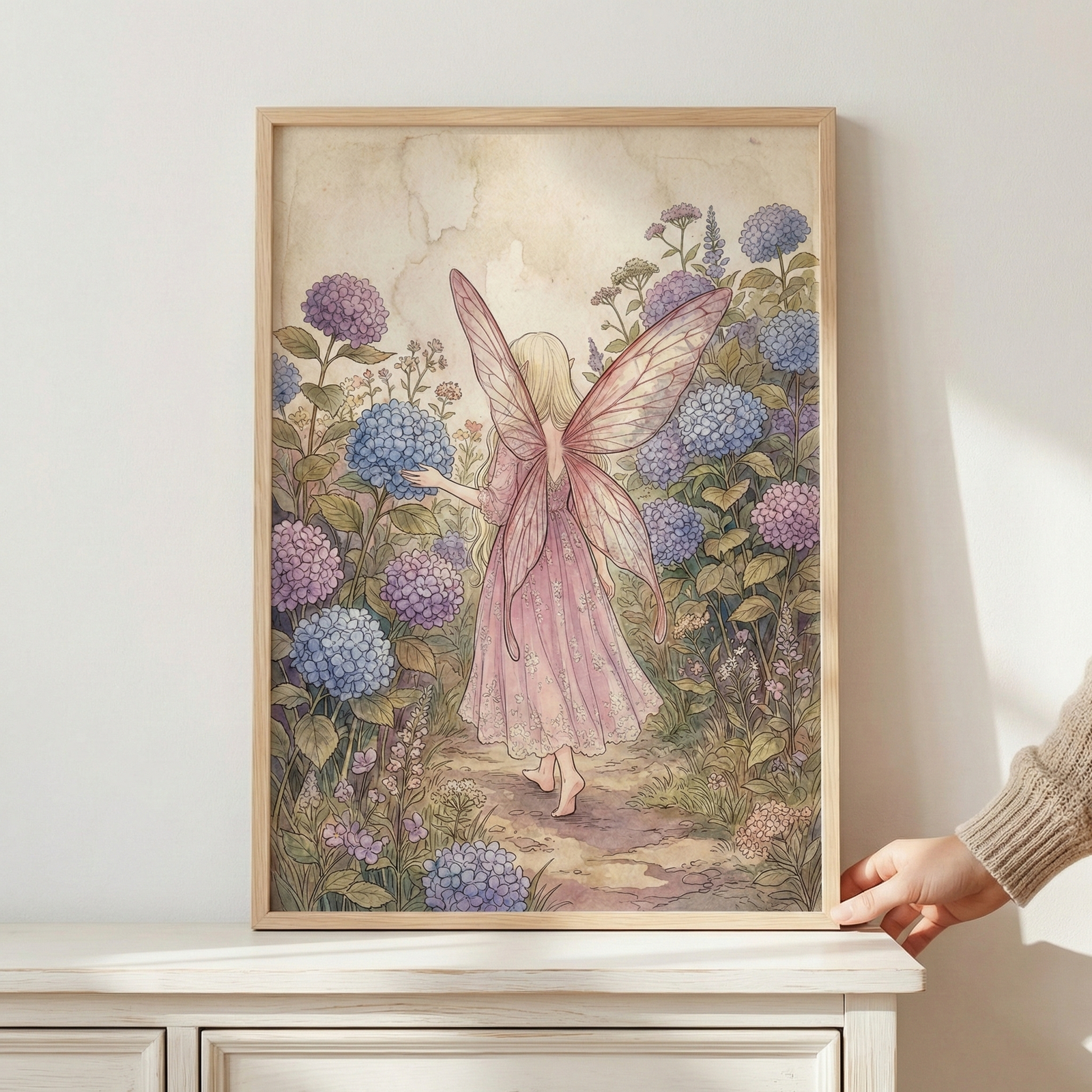A vertical framed illustration depicts a whimsical fairy from behind as she walks down a garden path. She has pale hair and large, translucent pink wings, wearing a delicate pink dress. Surrounding her are clusters of blue and purple hydrangea-like flowers. The piece features a soft, muted aesthetic with gentle brushstrokes. A hand holds the light wood frame resting on a white wooden surface.