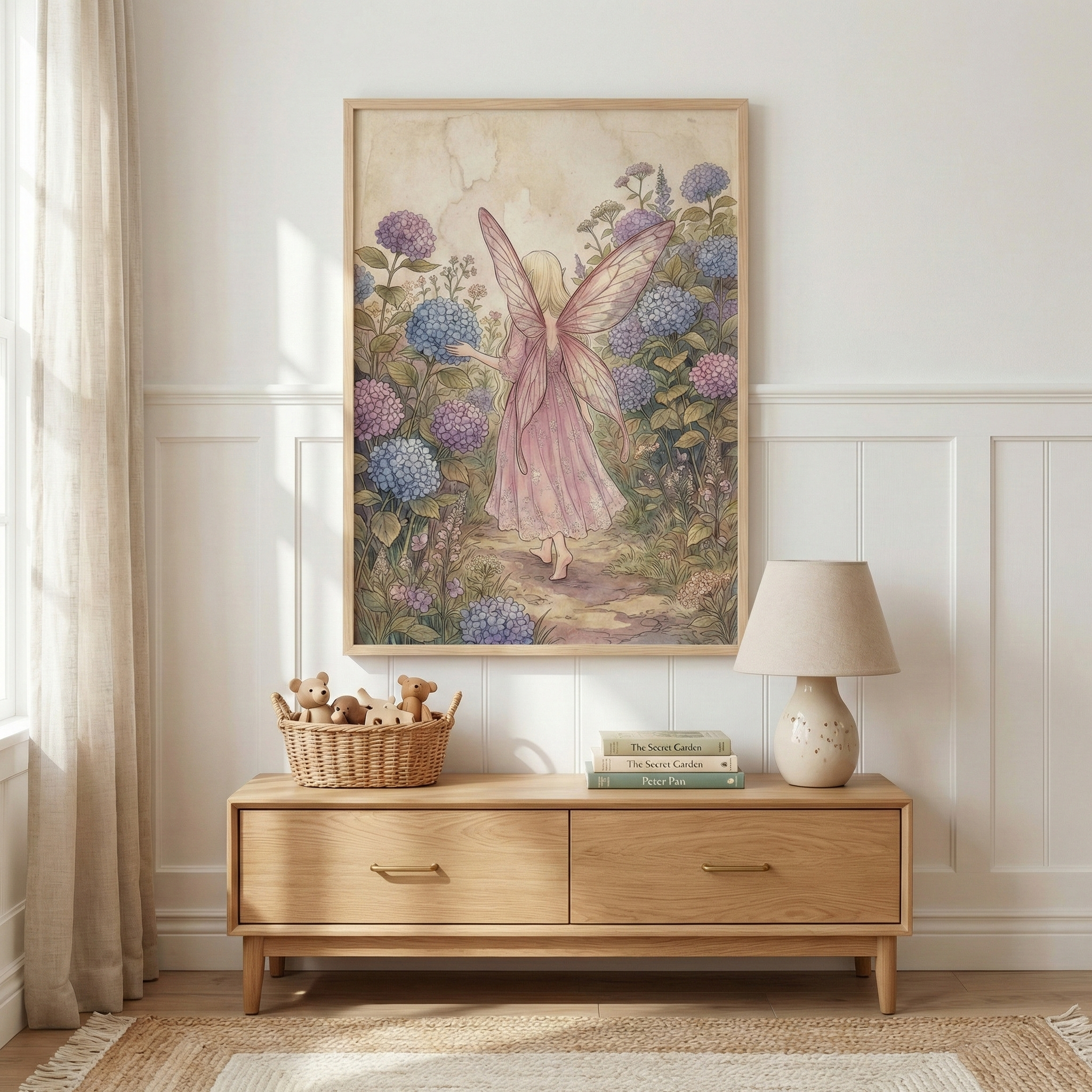 A whimsical nursery setting features a framed illustration hanging on a white paneled wall. The artwork depicts a winged figure in a pink dress walking through a lush garden of purple and blue hydrangeas. Below the artwork, a light wood two-drawer dresser holds a small woven basket filled with soft toy bears, a stack of books, and a cream ceramic lamp. A natural fiber rug and a neutral curtain complete the soft, warm aesthetic of the room.